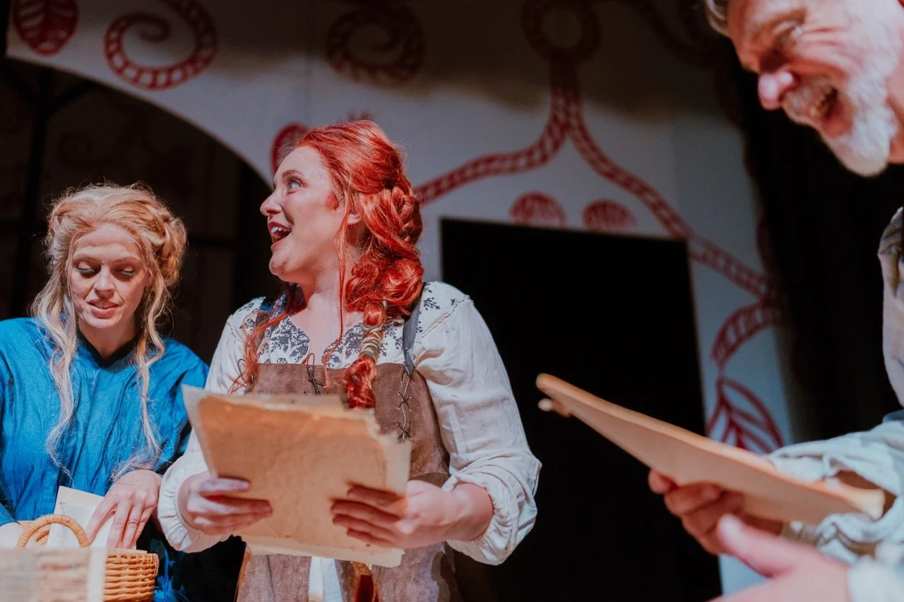 A theatrical scene with three actors in period costumes; one holds papers, another a basket.