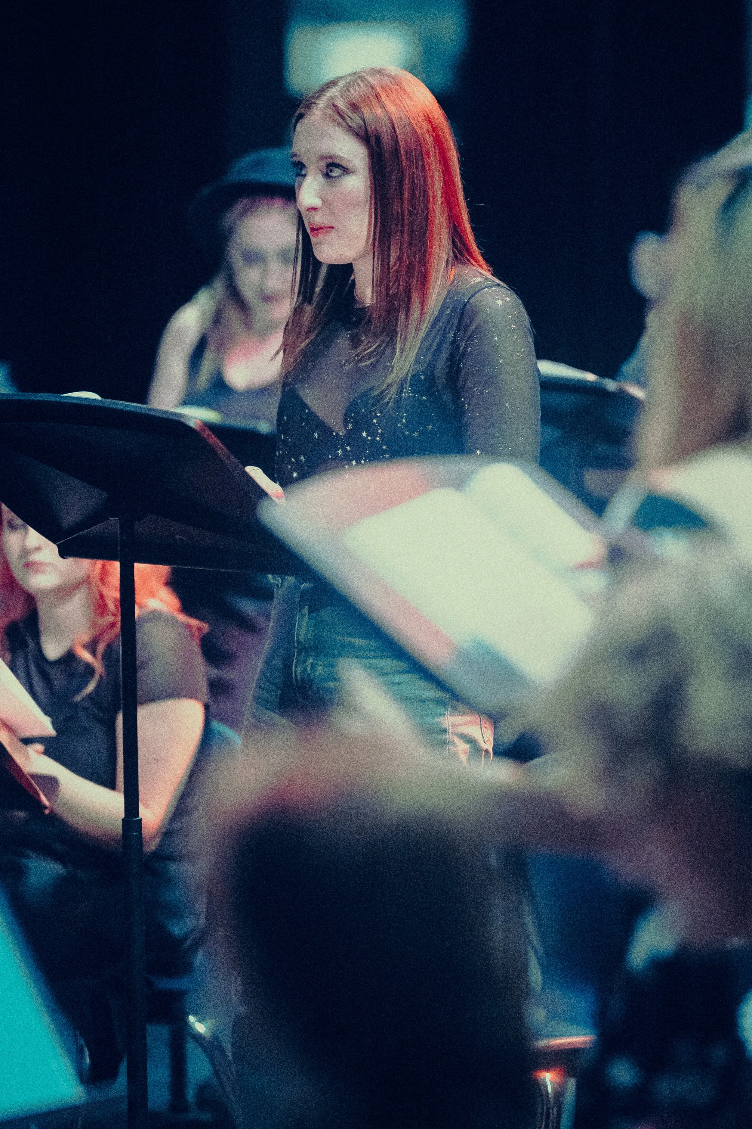 Woman singing in a choir with music stands, dark background, others in the background, stage lighting.