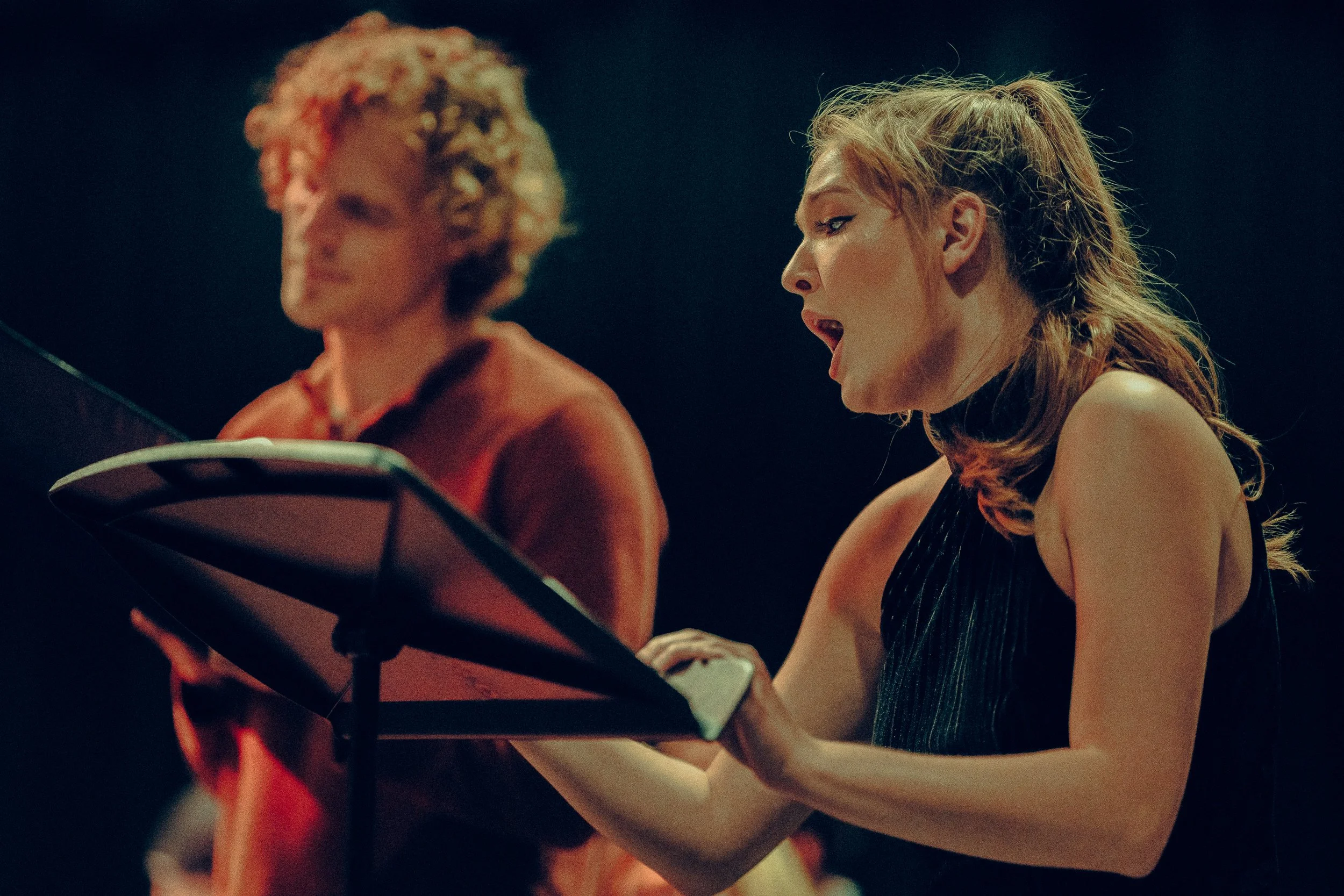A woman singing passionately into a microphone during a performance, with another person and music stand blurred in the background.