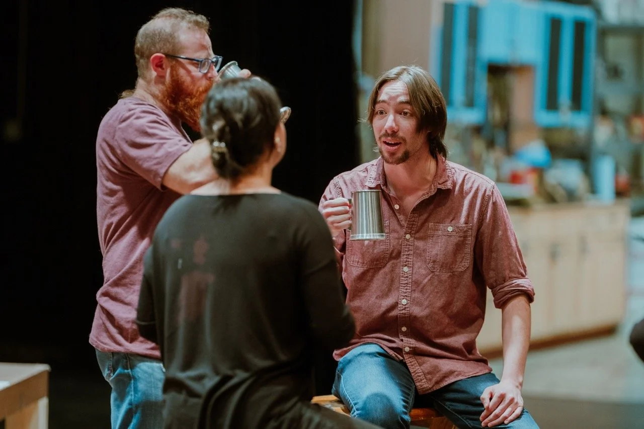 Three people socializing indoors, holding mugs, one with a beard drinking, another speaking, and a third with back turned.