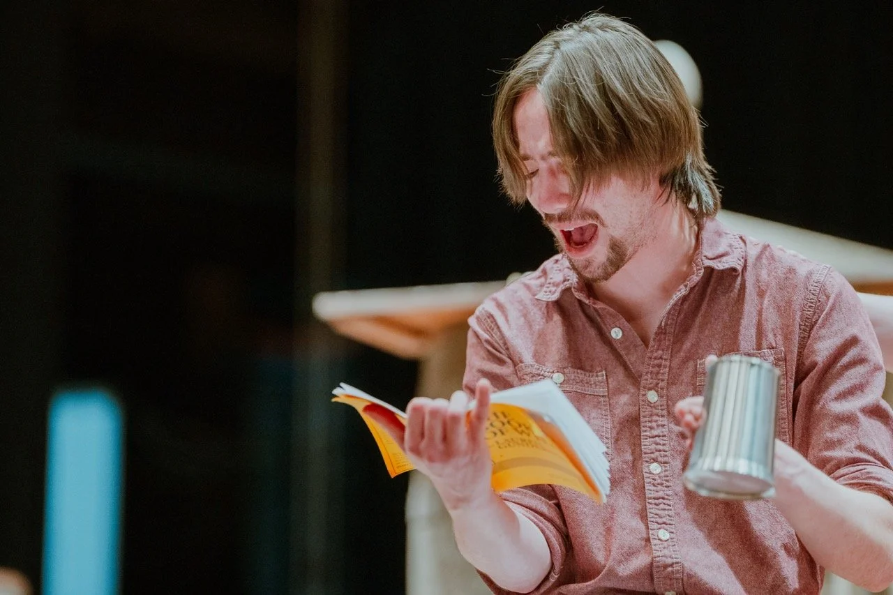 Person in a red shirt performing loudly with a book and a mug in their hands.