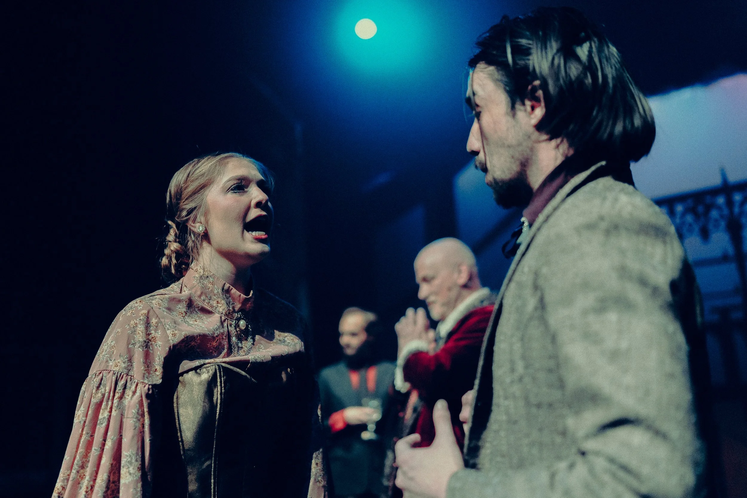 A theatrical scene with actors in period costumes performing on stage. The foreground shows a woman speaking passionately while a man listens. Background features other performers engaged in a scene, set under dramatic lighting.