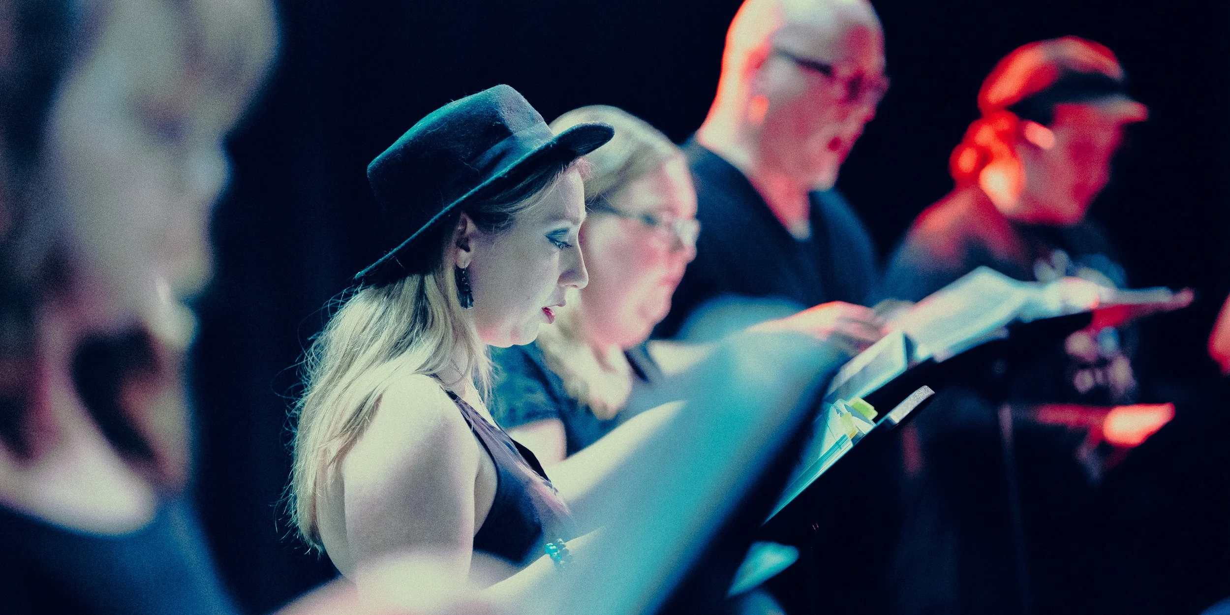 Group of people singing with music sheets under stage lighting.