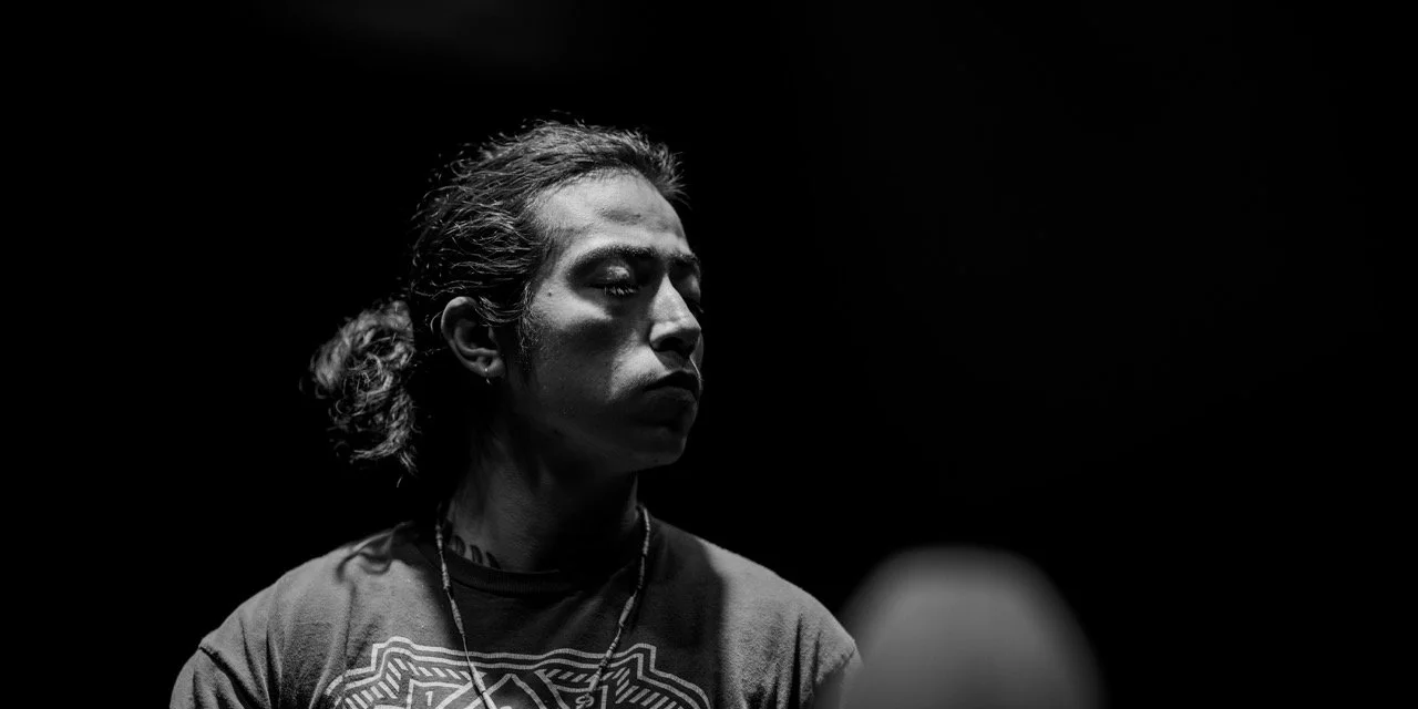 Black and white portrait of a person with long hair tied back, wearing a patterned shirt, in a dimly lit setting.