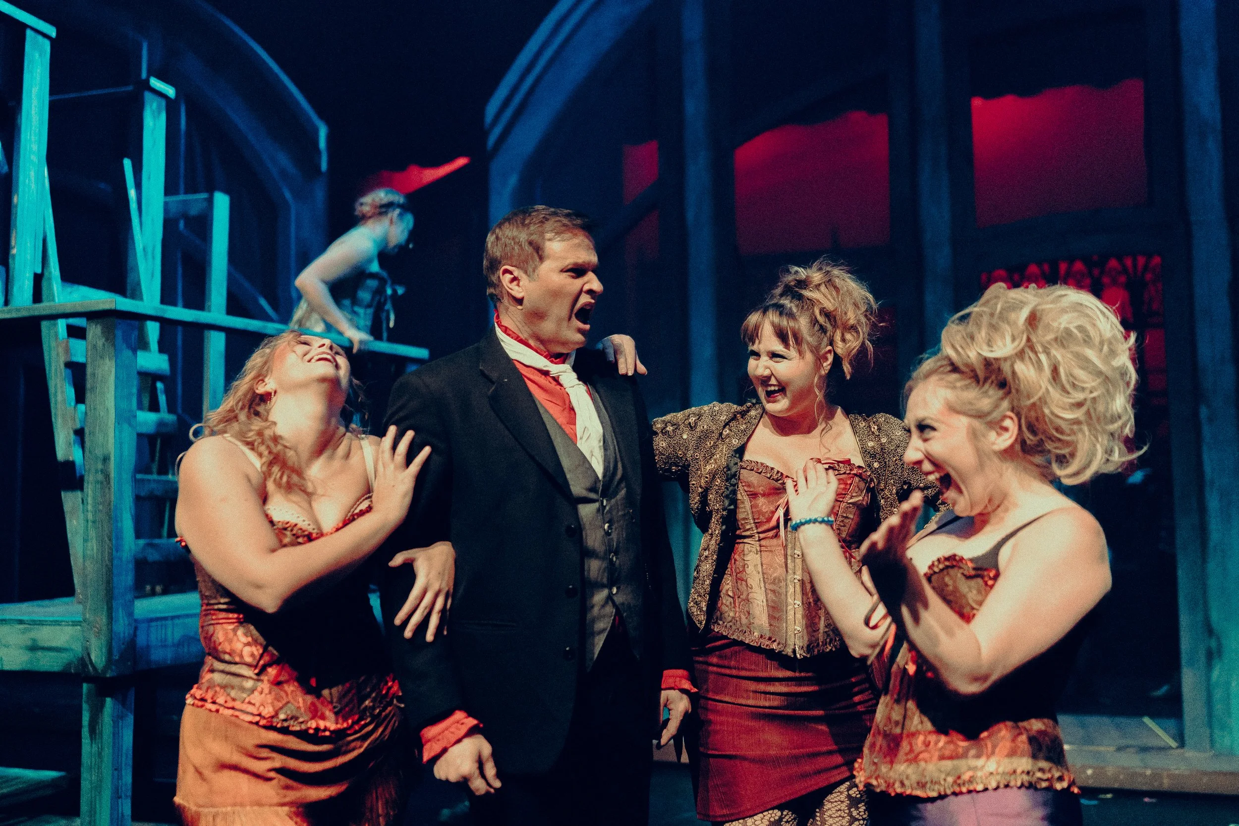 Group of actors in theatrical costumes laughing together on stage in colorful lighting and set.
