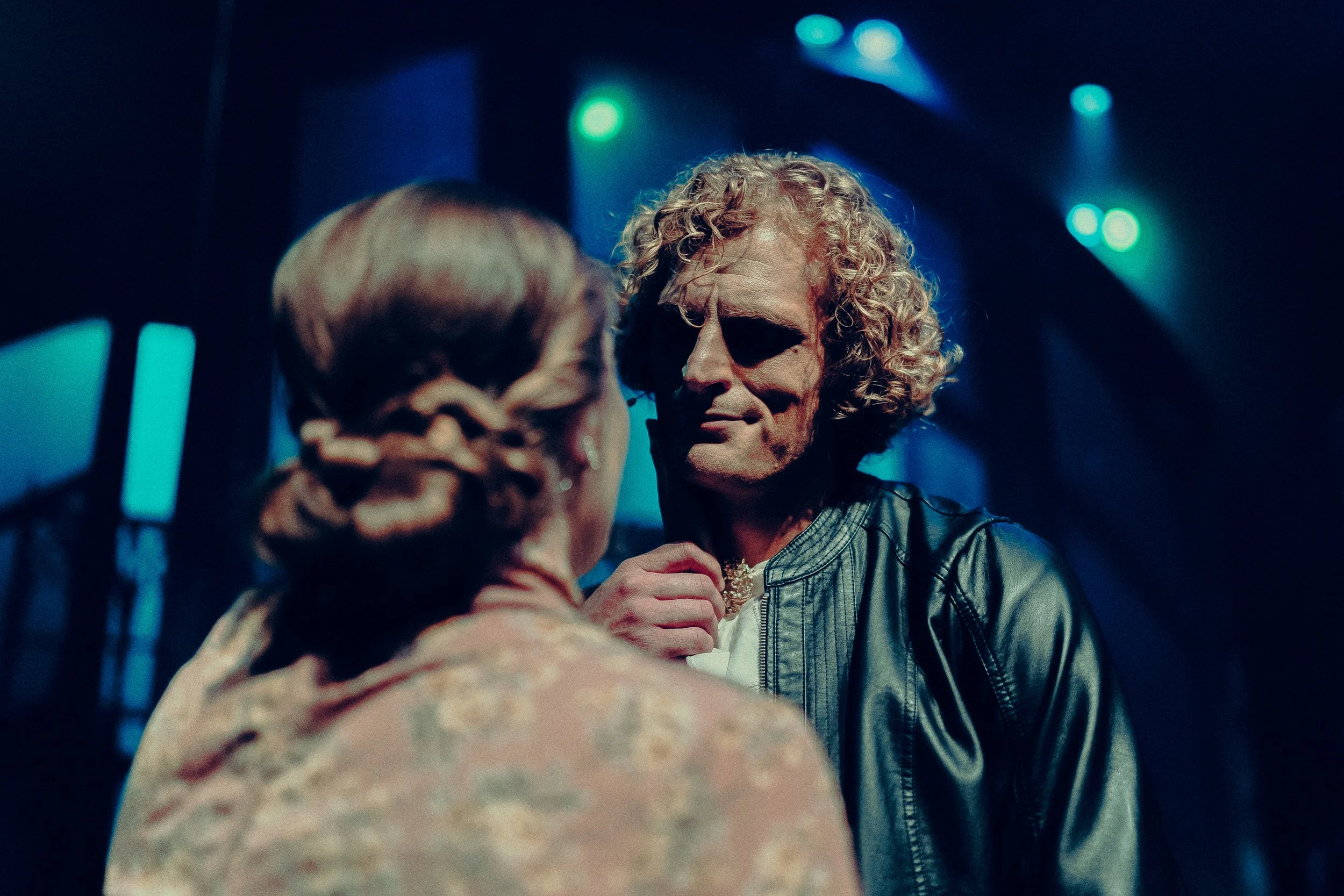 A person with curly hair in a leather jacket facing a person with a floral-patterned shirt in a dimly lit setting with blue and green lights.