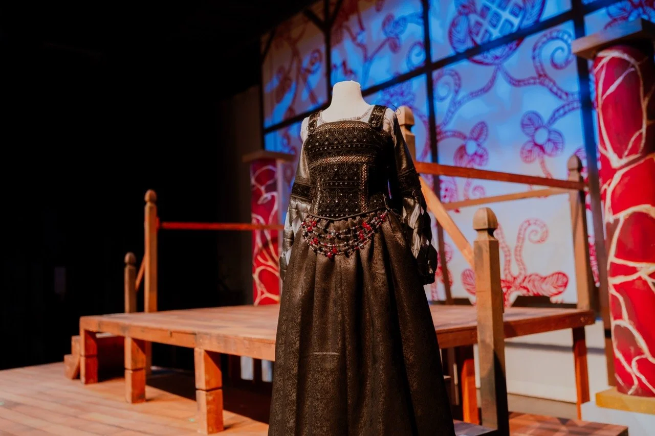 Elegant black dress with intricate detailing on mannequin in theater set with wooden stage and decorative backdrop.