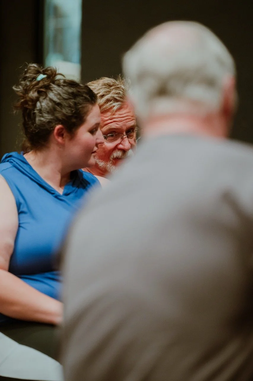 A group of people engaged in conversation in a casual indoor setting, with one individual wearing a blue shirt and another in focus with glasses.
