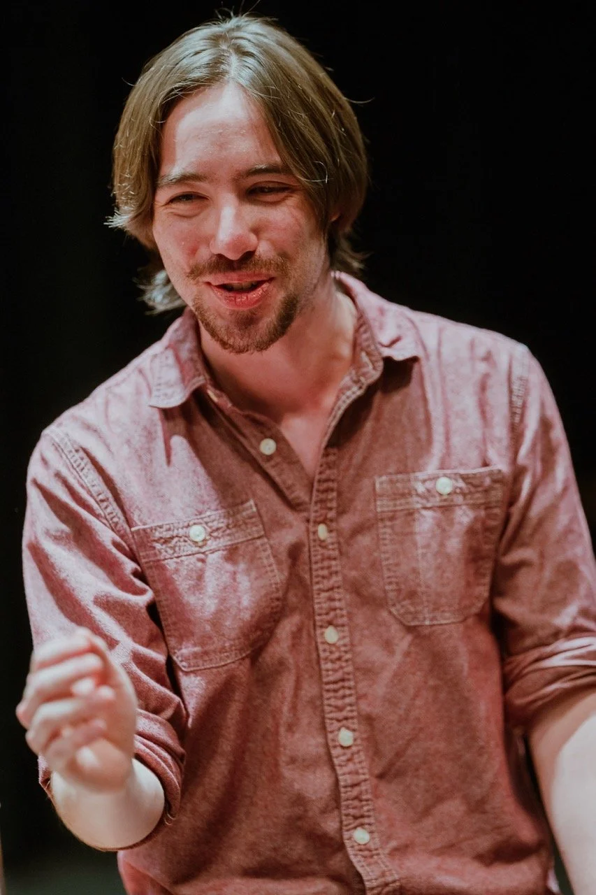 A person with shoulder-length hair and facial hair, wearing a red button-up shirt, speaking with an expressive gesture.