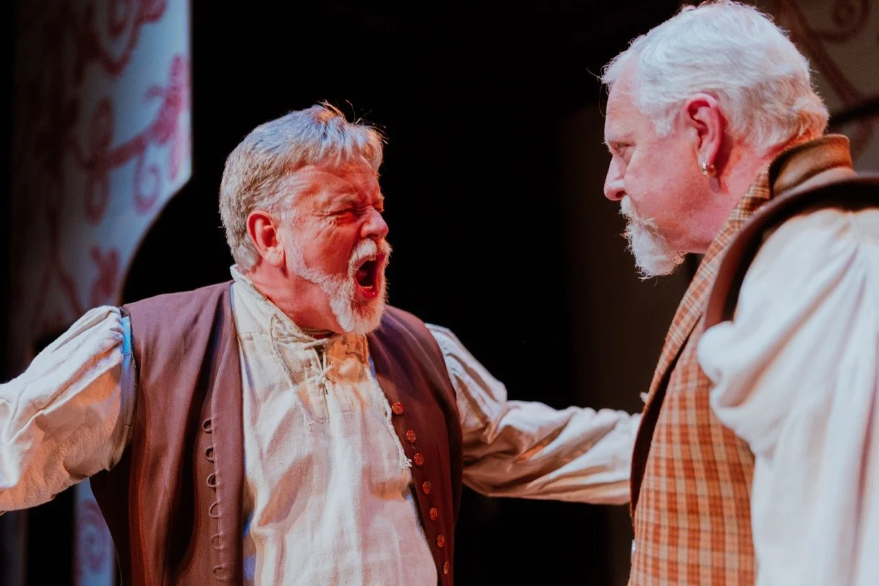 Two older men in historical costumes, engaged in a dramatic conversation on stage during a theater performance.