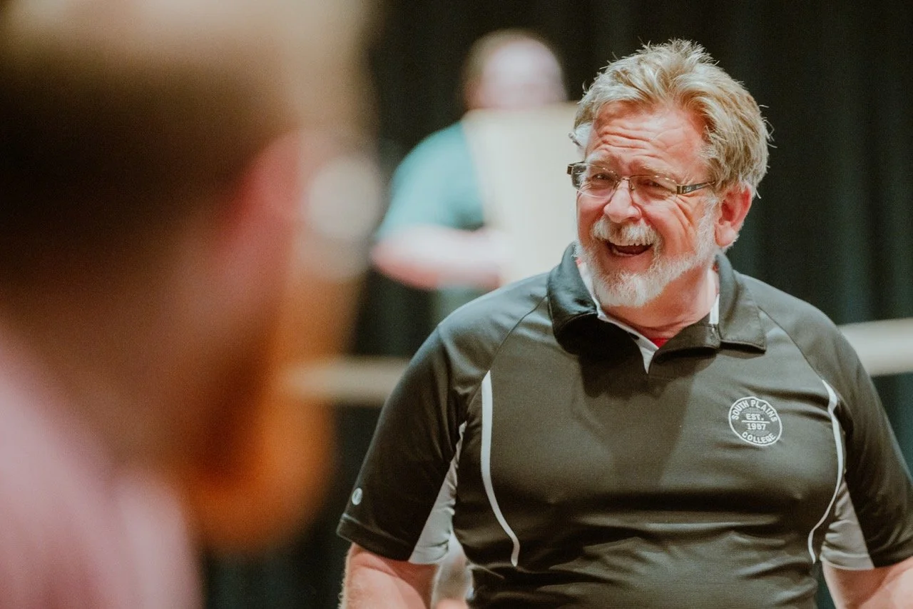 Laughing man wearing glasses and a black polo shirt, indoors.