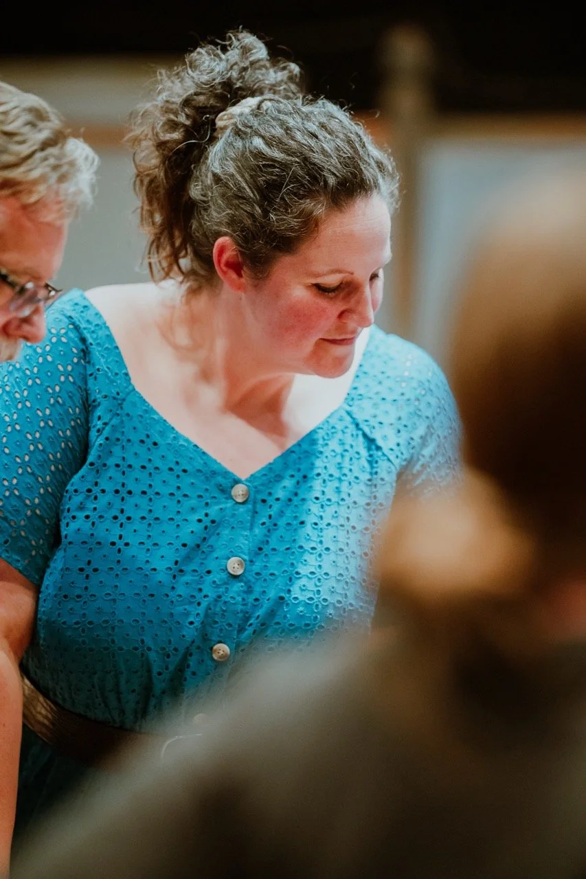 A woman in a blue dress with a textured pattern, looking down, with her hair pulled back, in a casual indoor setting.
