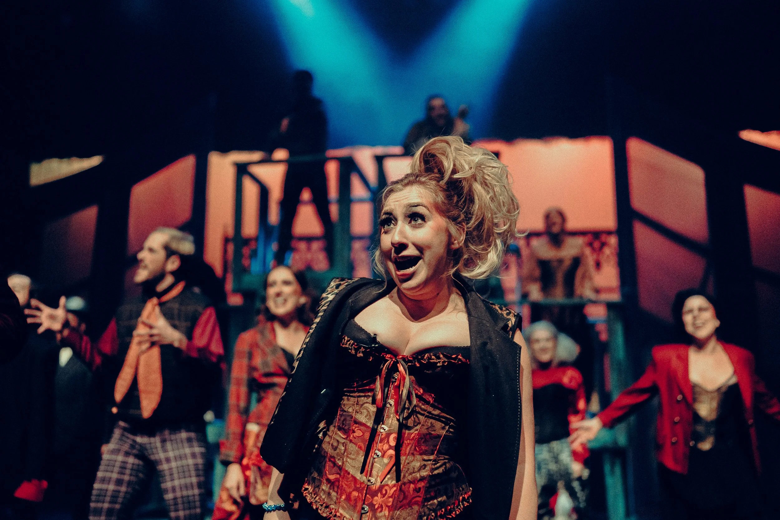 Stage performance with actors in colorful costumes, featuring a woman in a corset singing energetically under dramatic lighting.