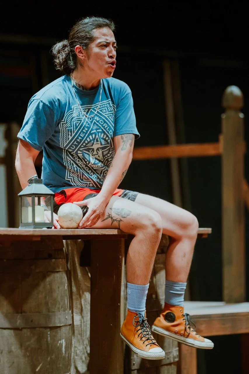 Person sitting on a wooden structure holding a prop skull, wearing a blue graphic T-shirt, orange high-top sneakers, and blue socks, with a lantern beside them.