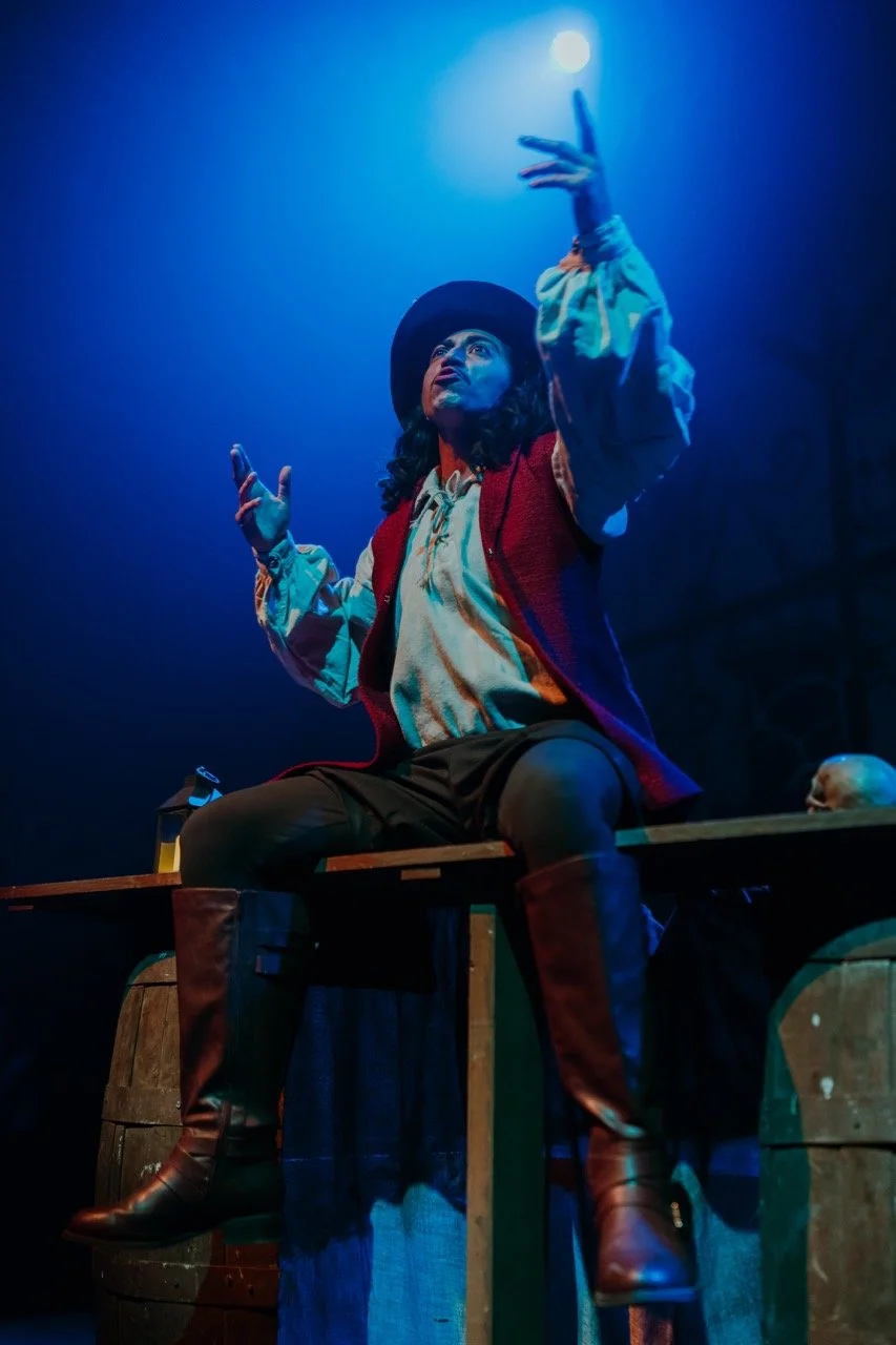 Person in theatrical costume sitting on a wooden beam with arms raised, wearing a hat and boots. Blue lighting creates a dramatic mood with a skull and lantern nearby.