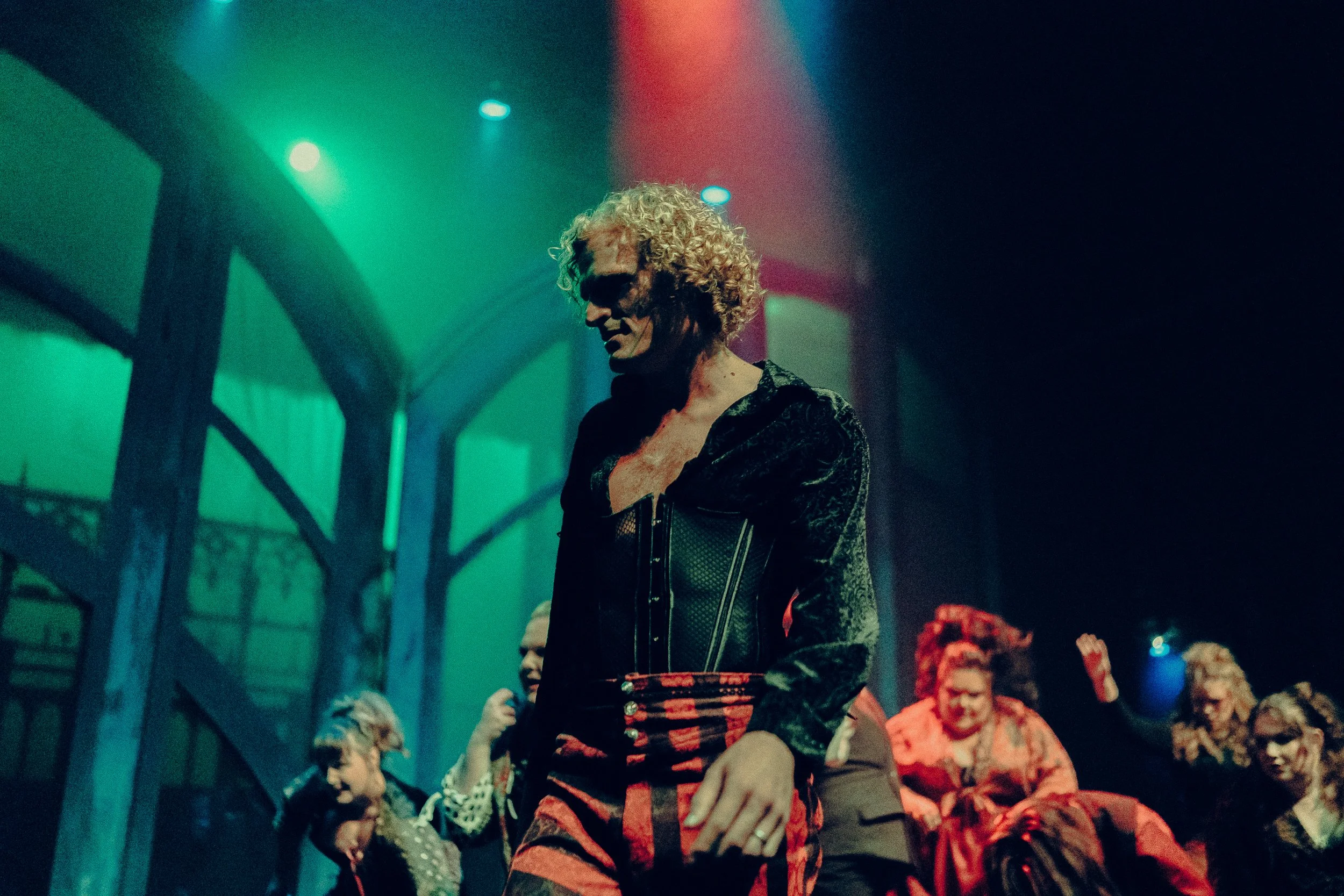 Stage performance with a man in a theatrical costume, featuring red and black attire, surrounded by other performers under colorful stage lighting.
