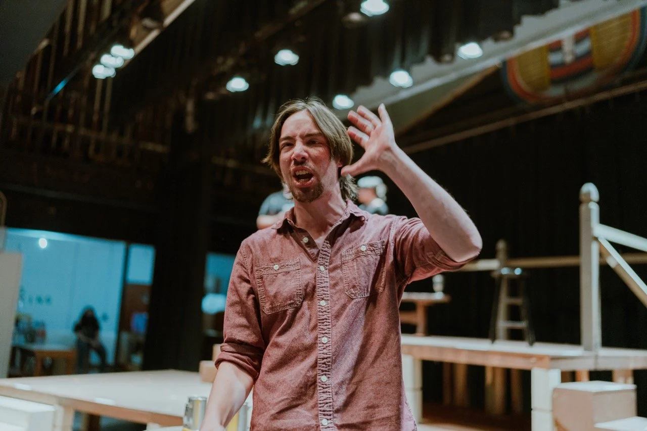 A person passionately speaking on stage with raised hand in a theater setting.