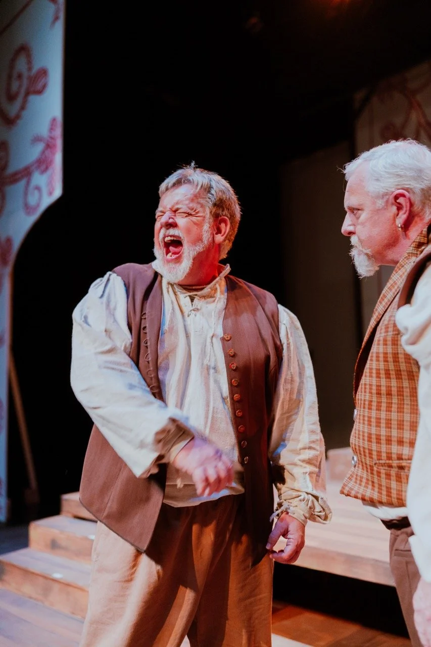 Two actors on stage wearing period costumes, one gesturing dramatically with an open mouth while the other watches.