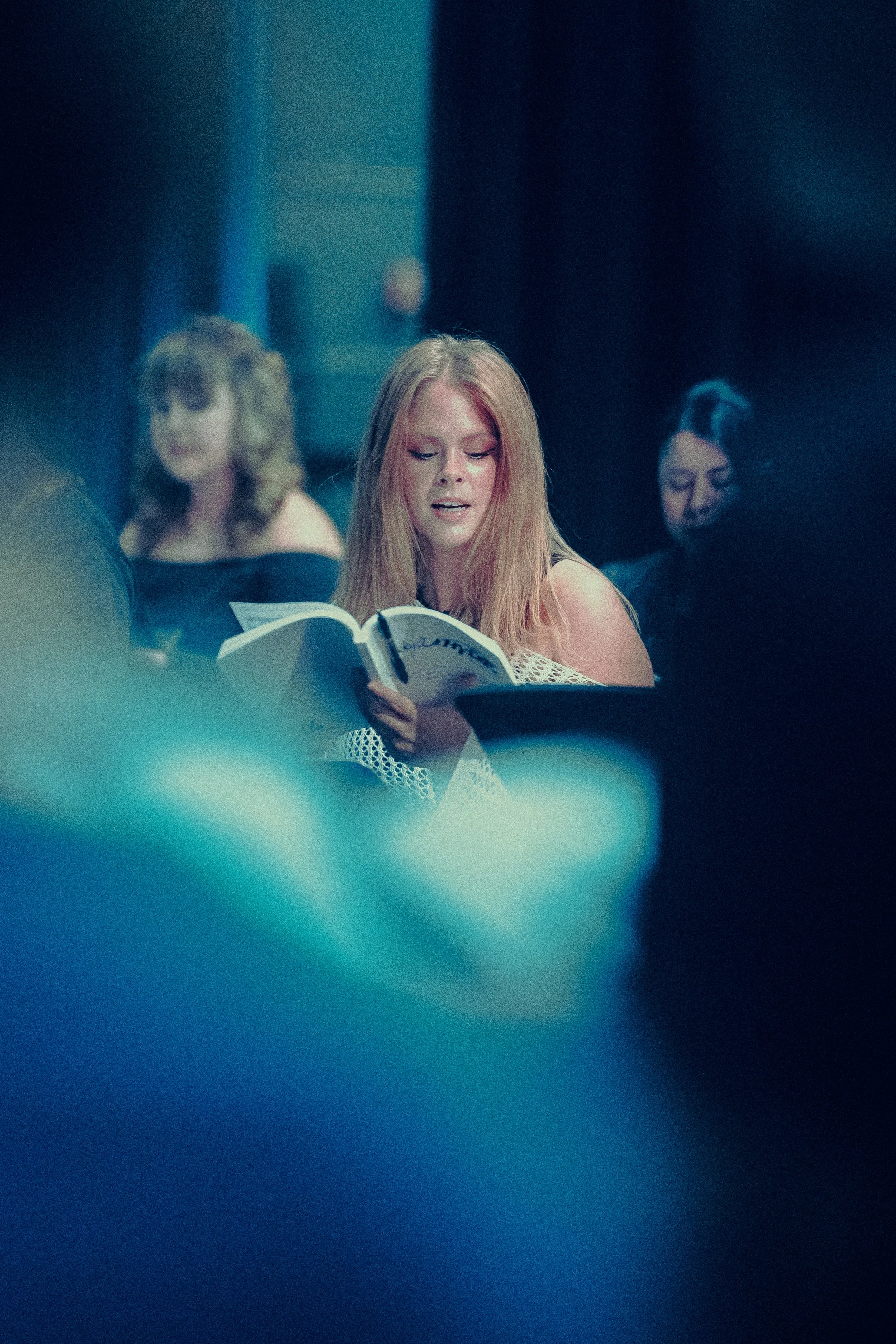 Woman reading a book in a dimly lit room, with people in the background.
