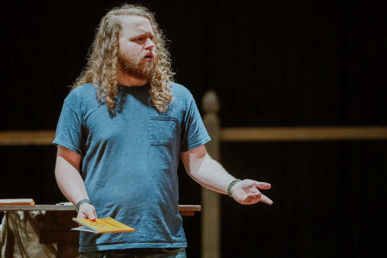 Man with long curly hair, beard, and blue shirt holding a pamphlet while gesturing with his other hand on a dark stage background.