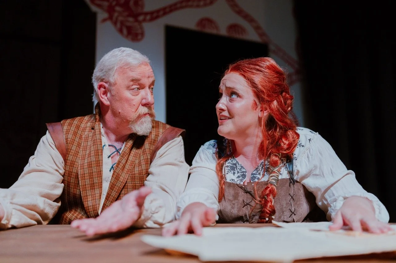 Two actors in period costumes, engaged in conversation at a table, one with gray hair and beard, the other with red braided hair, performing a scene.