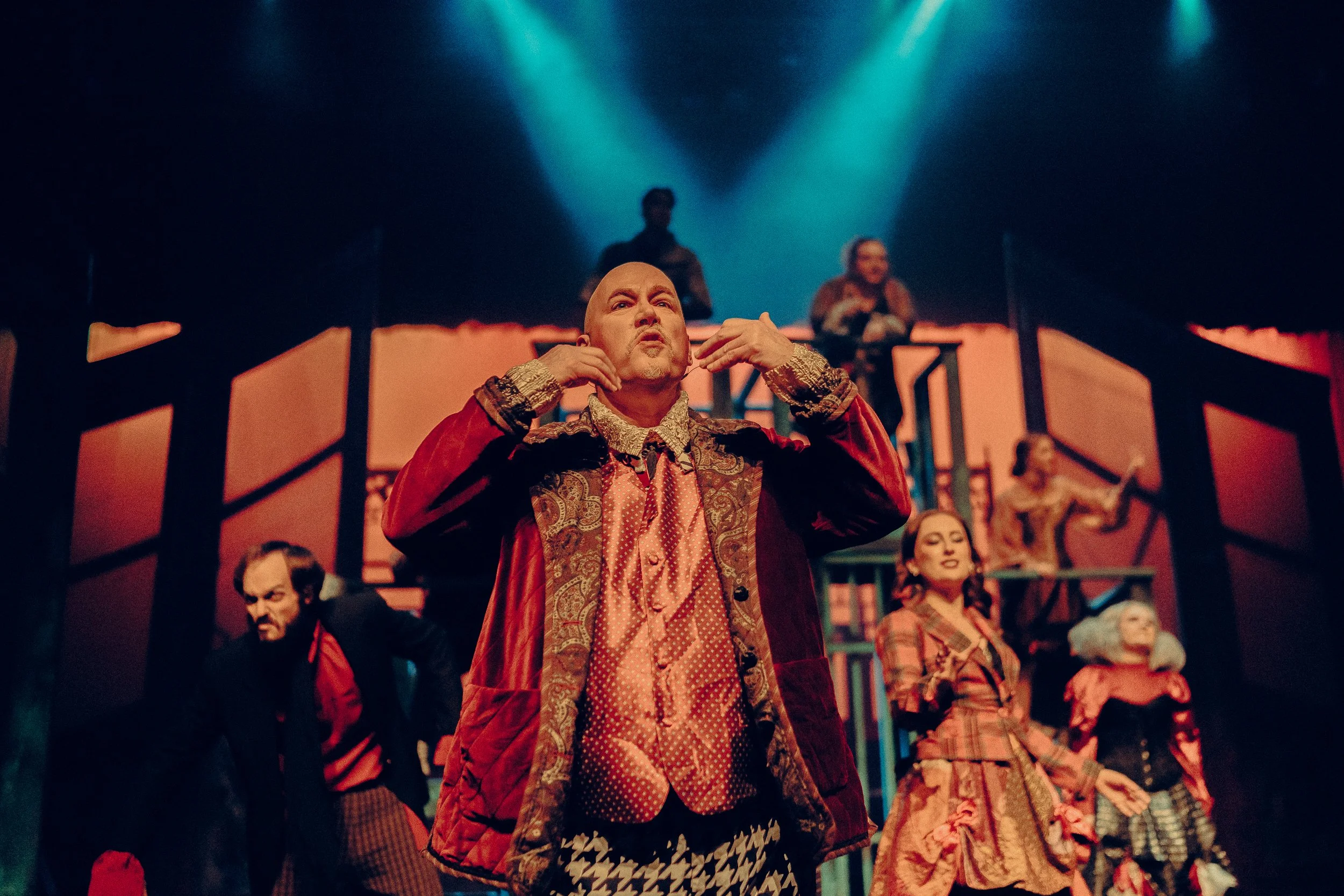 Theatrical performance with actors in colorful, vintage costumes on stage under dramatic lighting. A central figure in a red velvet coat gestures expressively, surrounded by other performers.