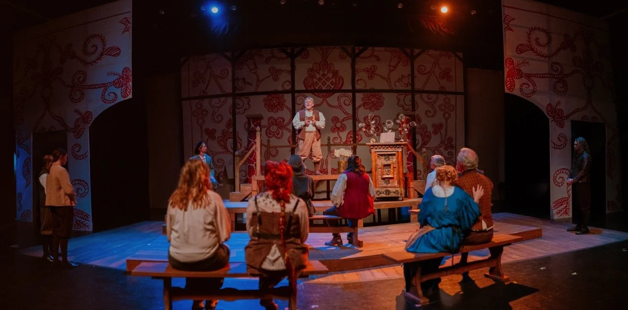 Theater scene with actors in period costumes on stage, featuring a central figure speaking or performing, surrounded by people seated on benches. The backdrop has floral patterns.