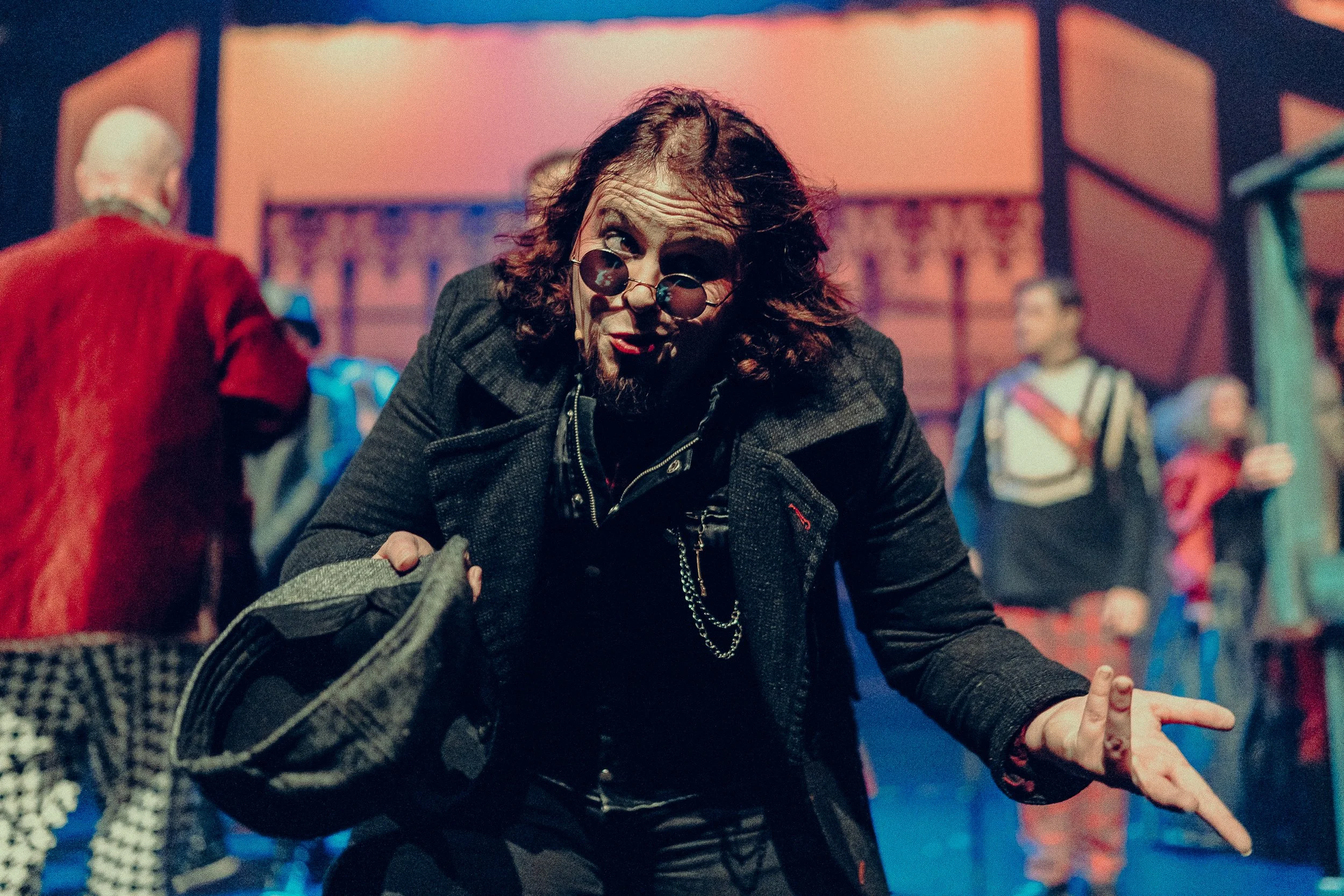 Person dressed in theatrical costume with glasses, holding a hat, and performing on stage with blurred background actors and dramatic lighting.