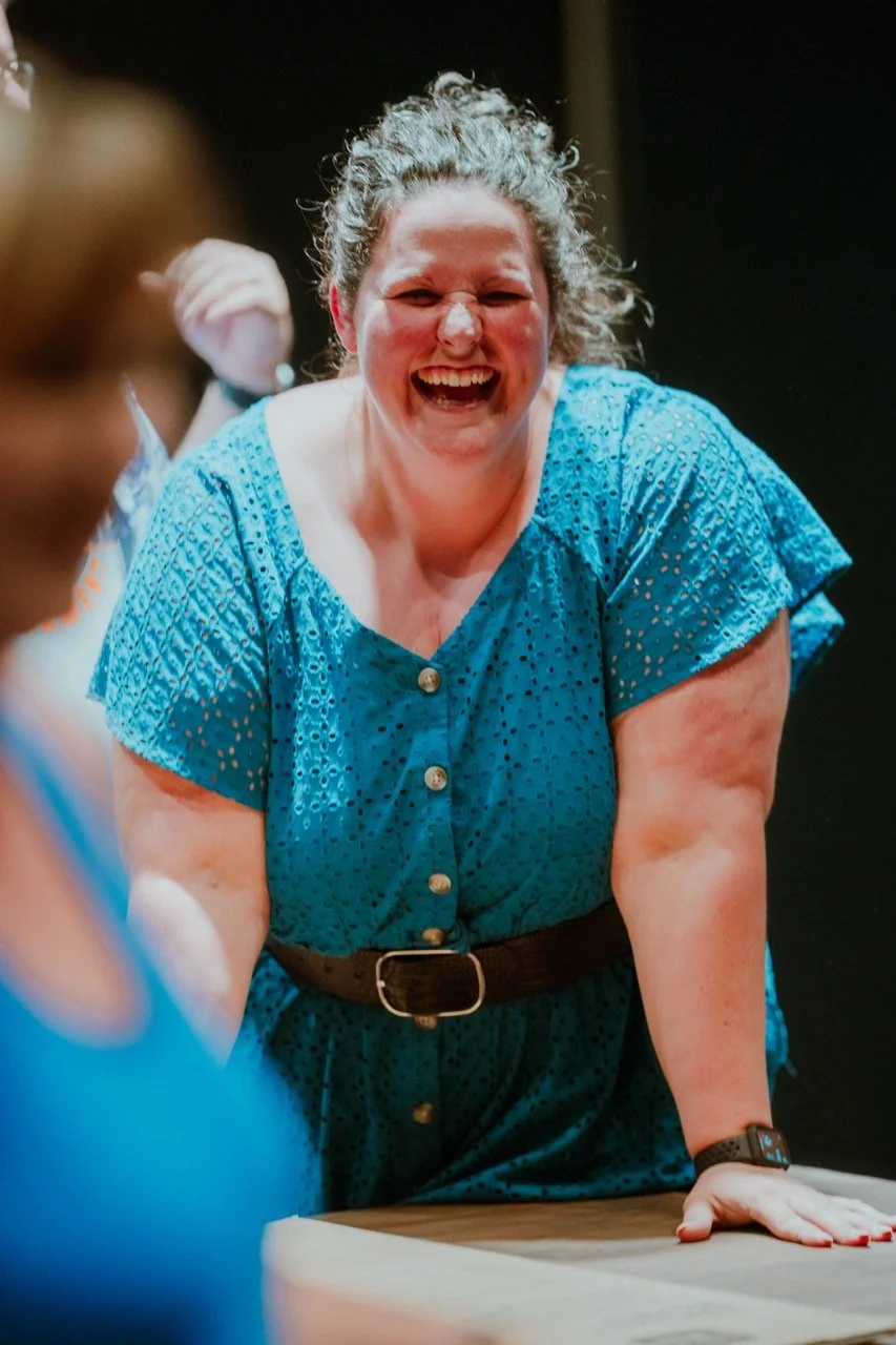 Woman in a blue dress laughing, leaning on a table, with people blurred in the foreground.