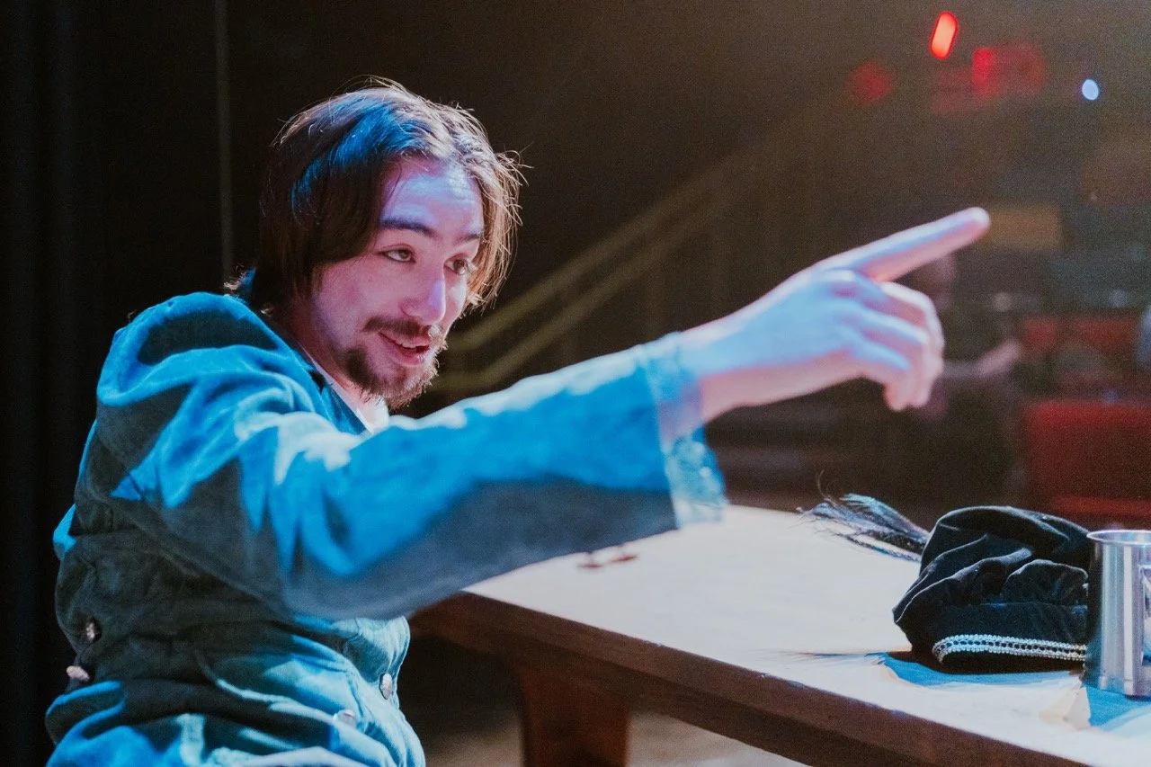 Man in blue costume sitting at a table on stage, pointing