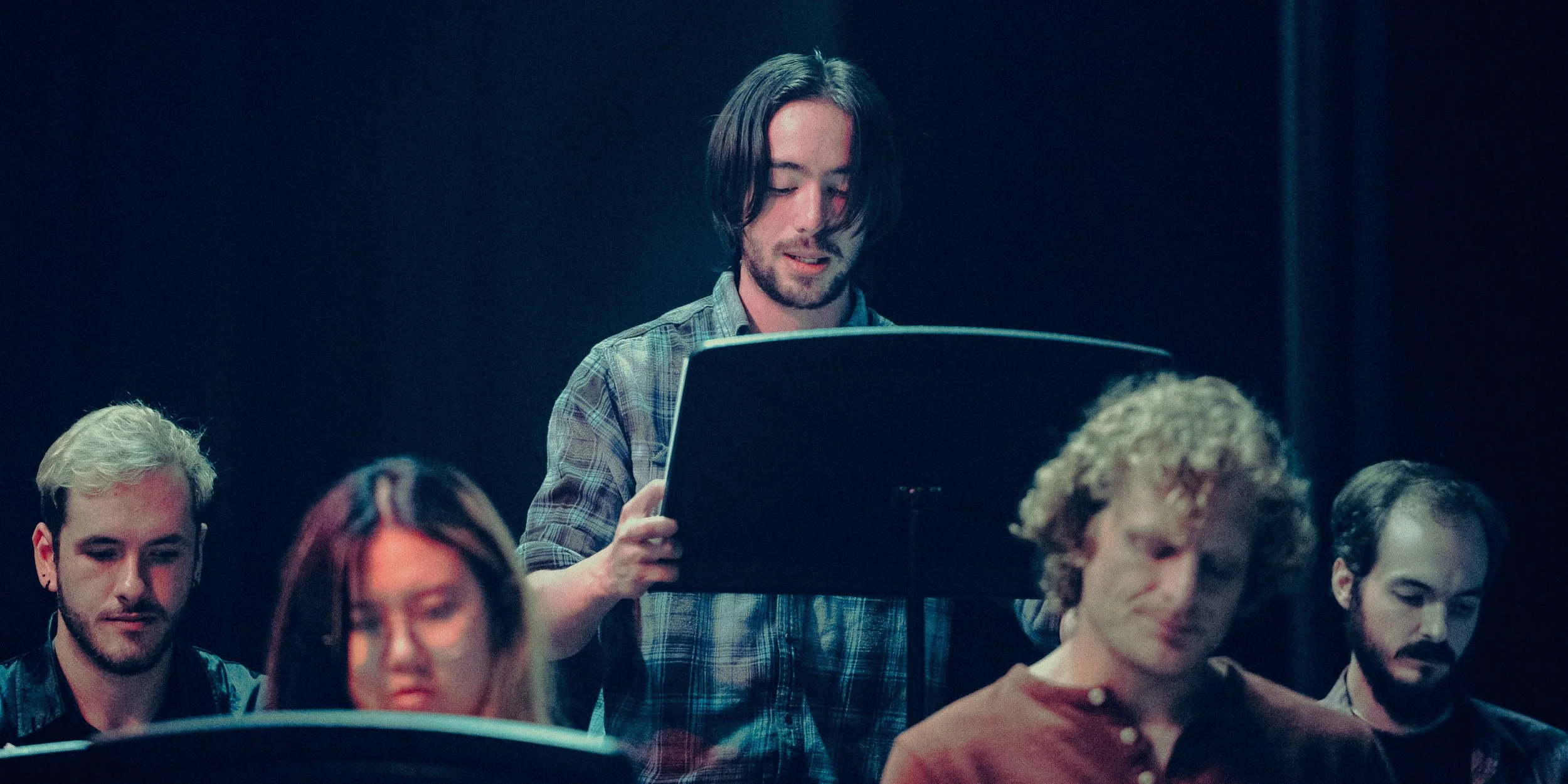 A group of people sitting and reading from music stands in a dimly lit room, with one person standing and reading at the center.