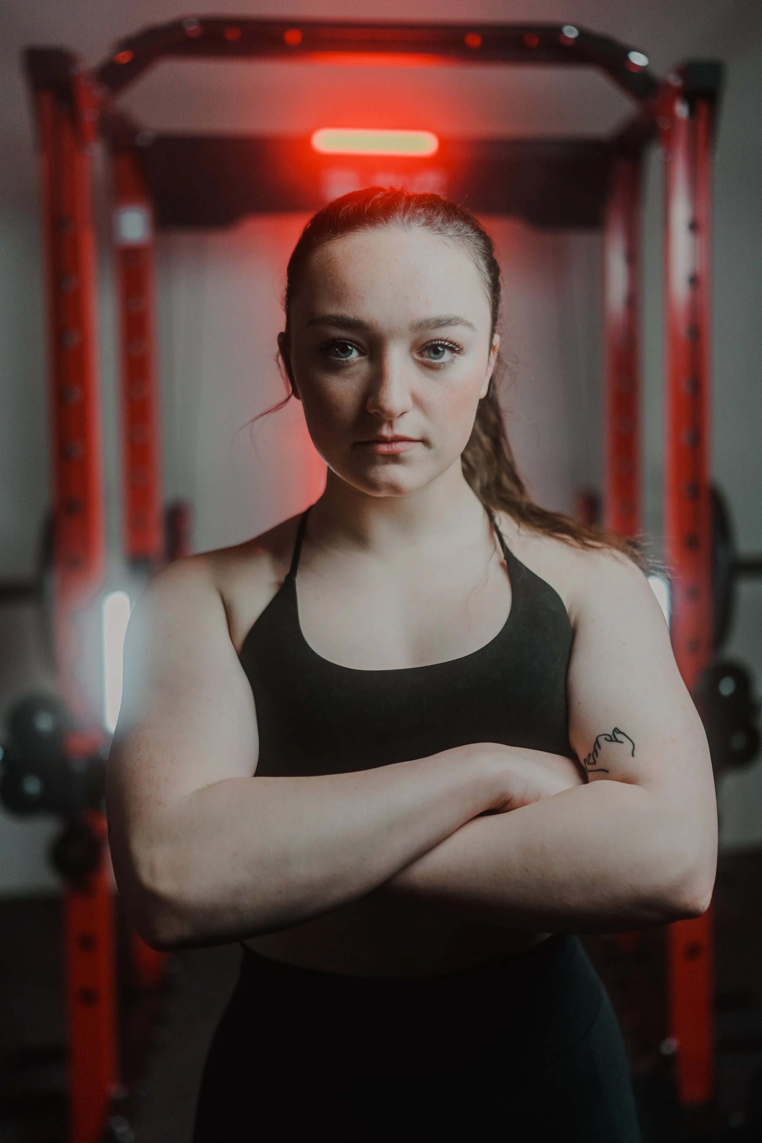 Person with arms crossed in front of gym equipment, wearing a black sports bra, with a tattoo on the arm, red lighting.