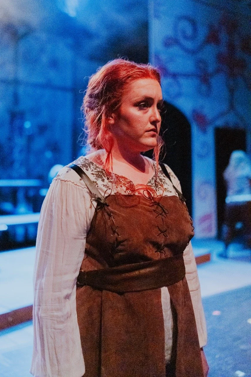 Person in medieval-style costume with reddish hair on stage, blue-hued lighting.