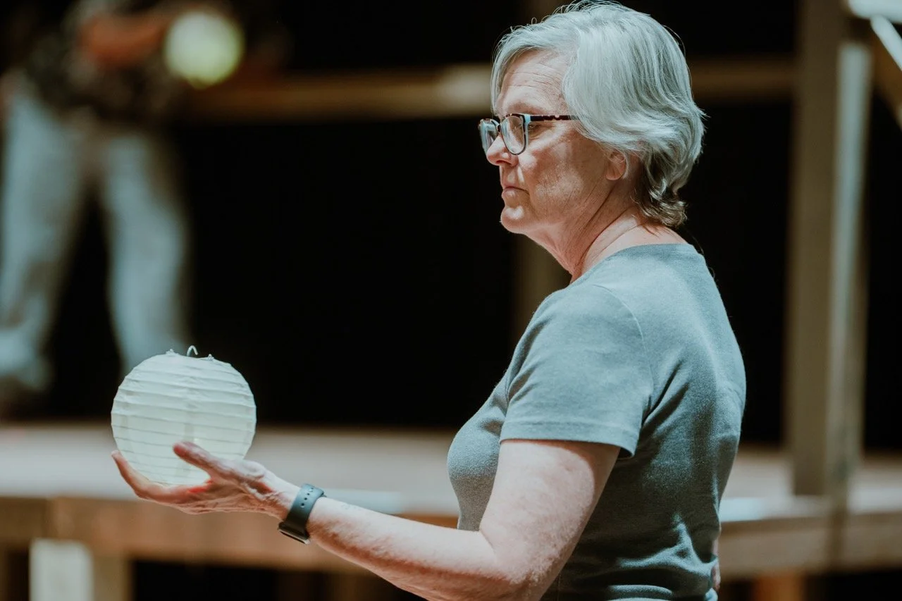 Elderly woman with glasses holding a paper lantern on a stage.