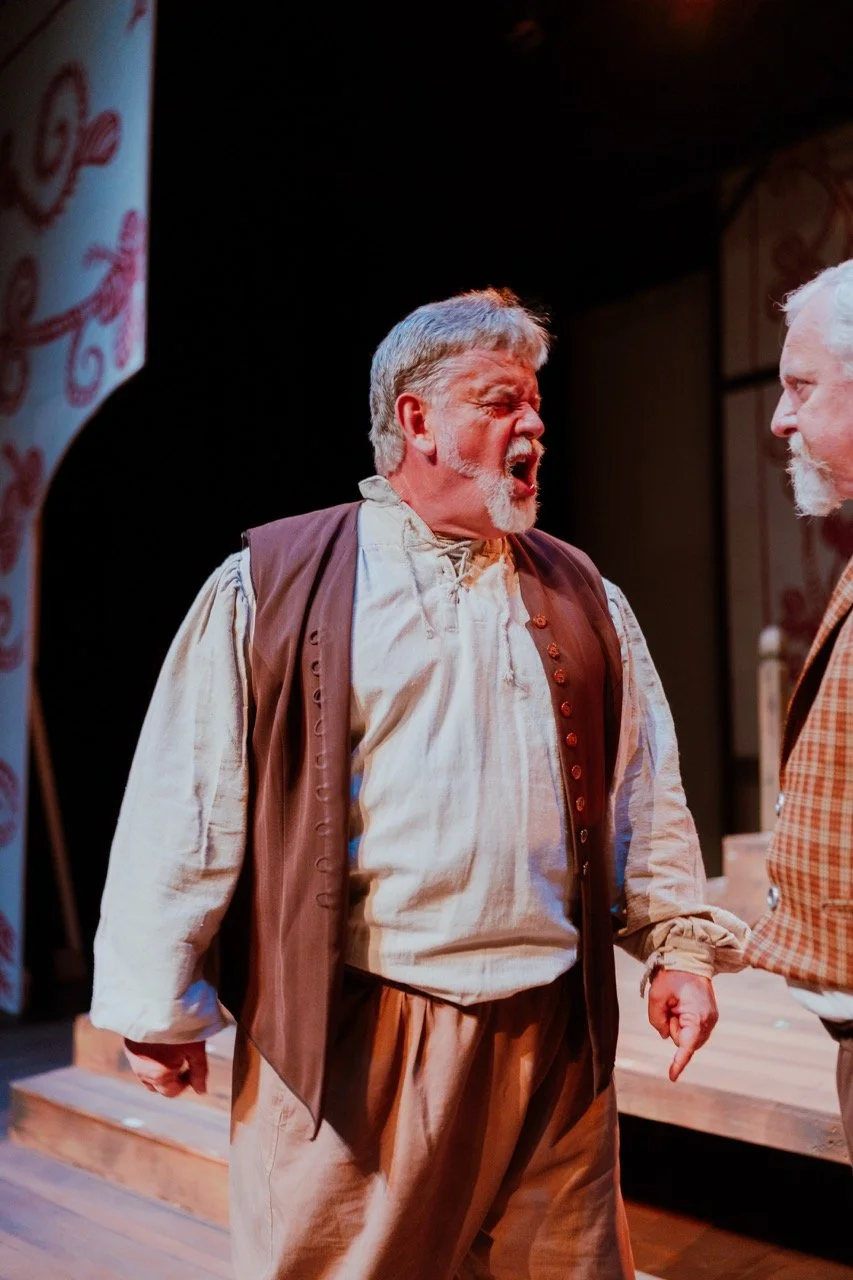 Older man in historical costume yelling on stage during a play, with another actor partially visible.