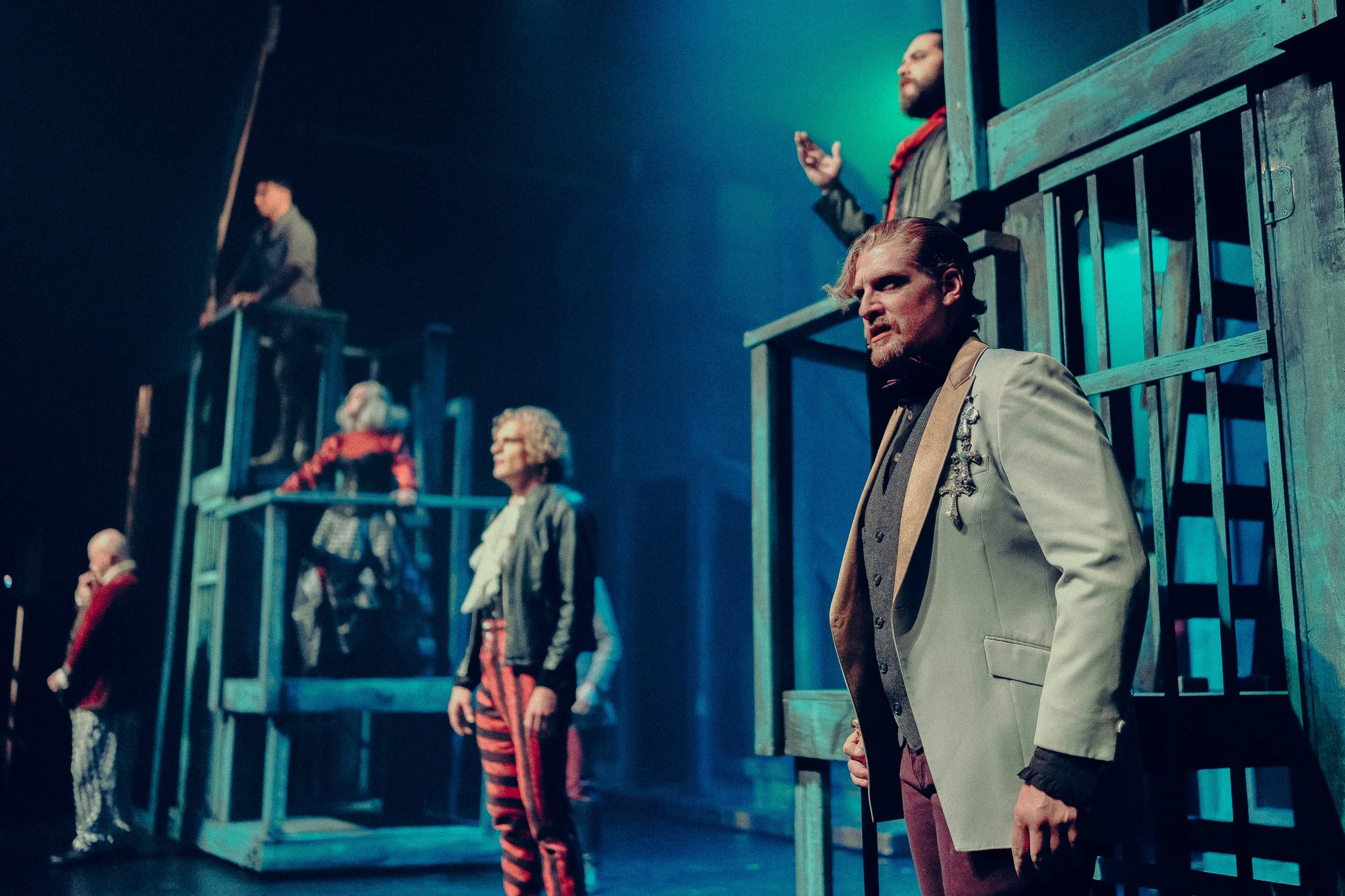 Theater performance with multiple actors on a layered set, under dramatic lighting. The scene includes actors in vintage costumes, standing and interacting on wooden scaffolding structures, conveying a dramatic or historical narrative.