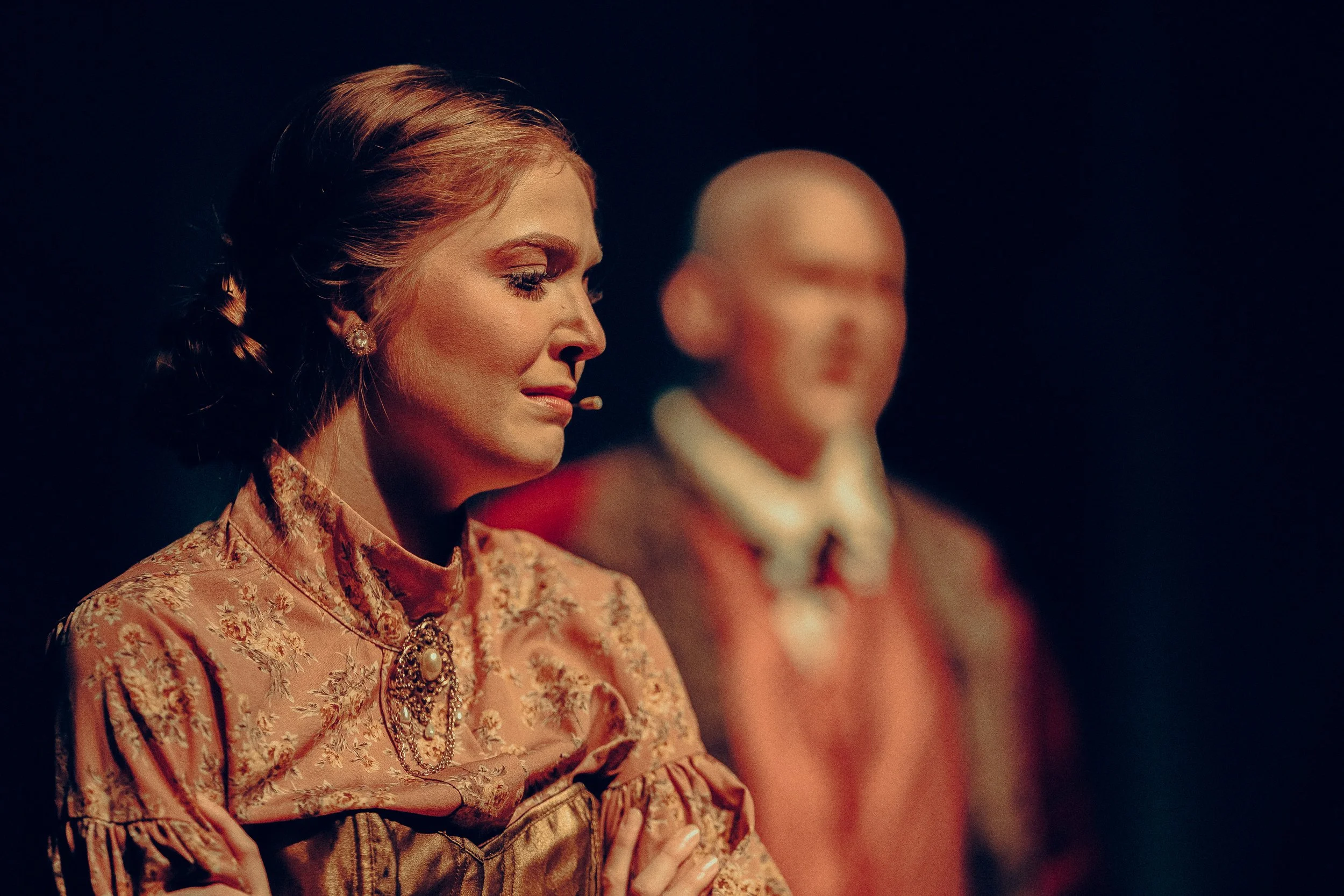 A woman in period costume on stage with a microphone, performing in a theater production.