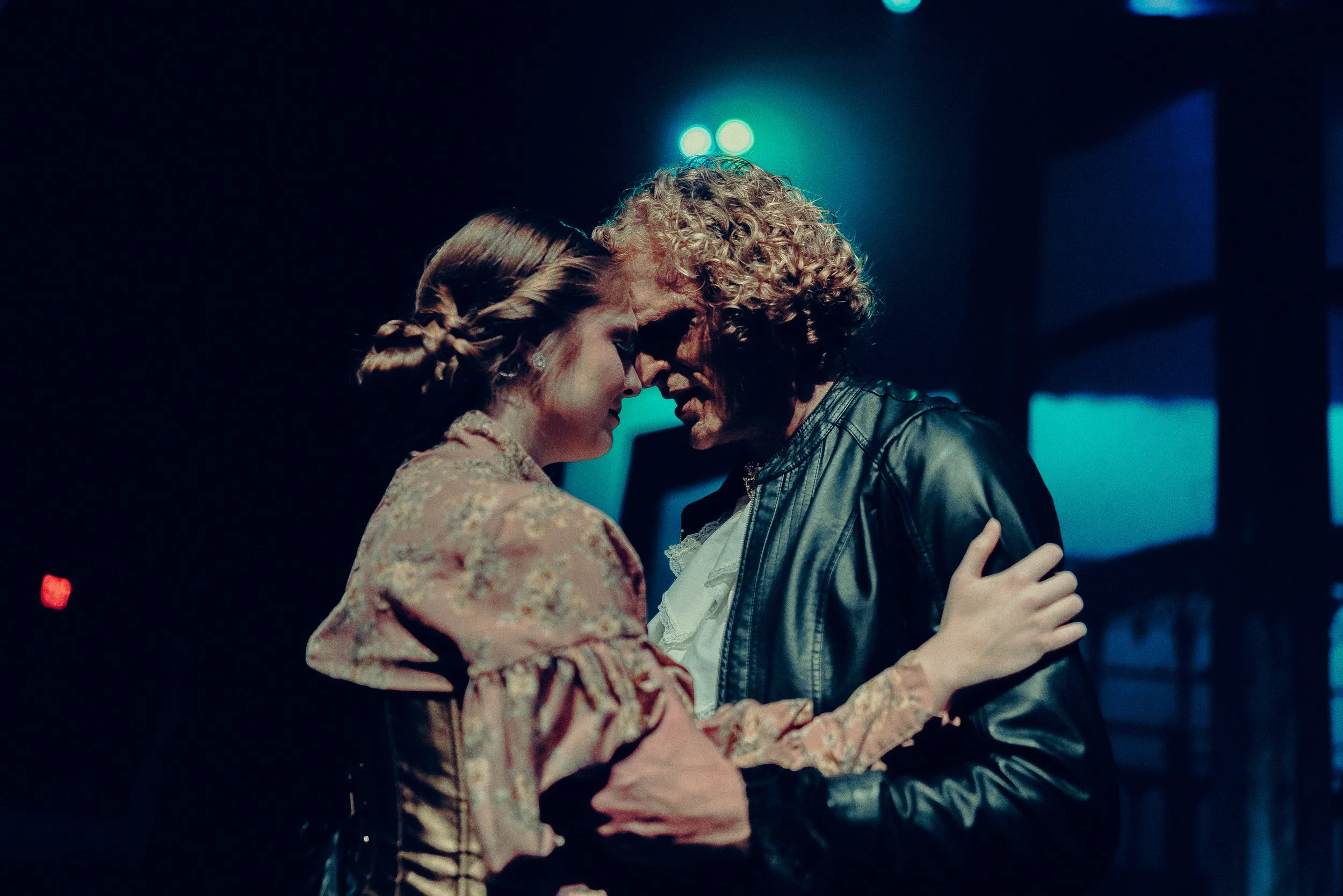 Two people in an intimate moment on stage, one wearing a floral costume, the other in a leather jacket, with blue lighting.