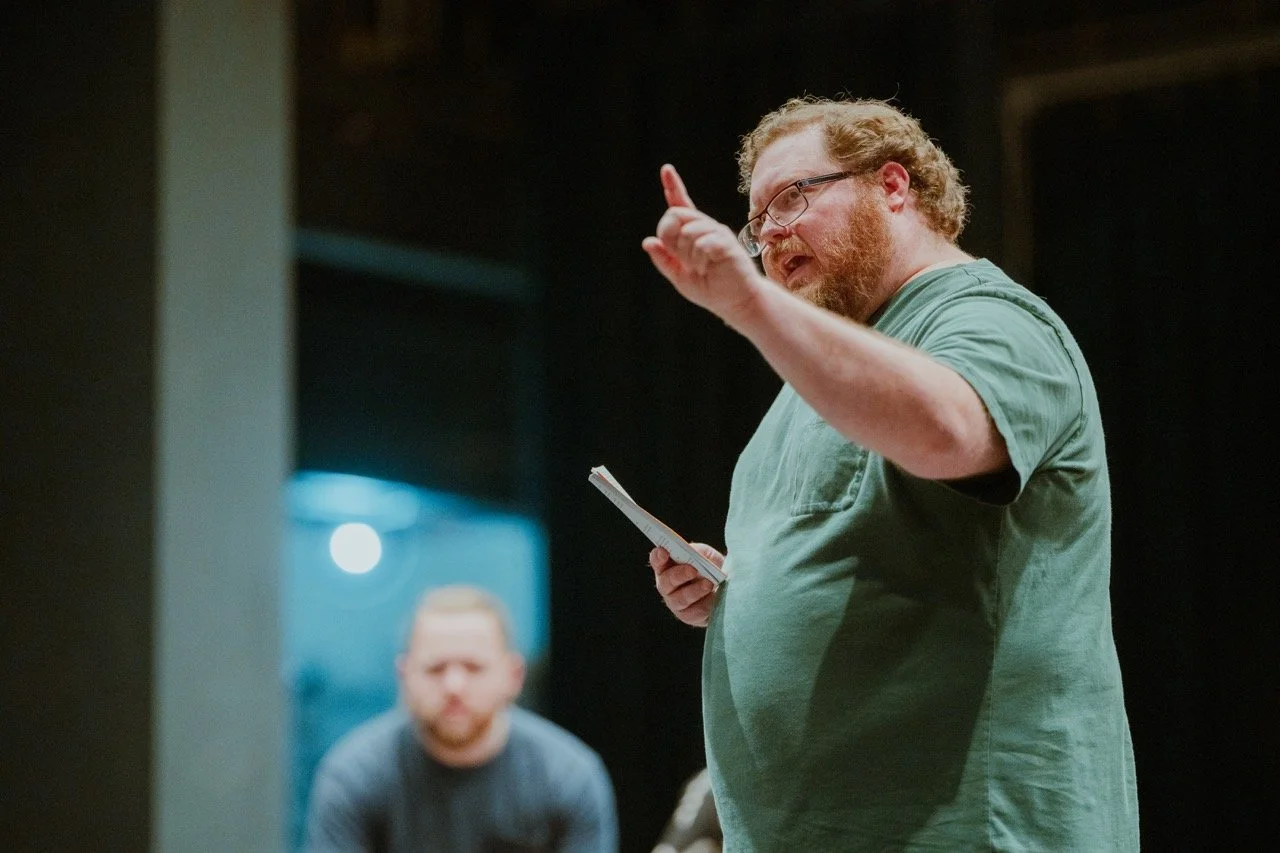 Man with glasses and beard holding paper, gesturing while speaking, with another person blurred in the background.