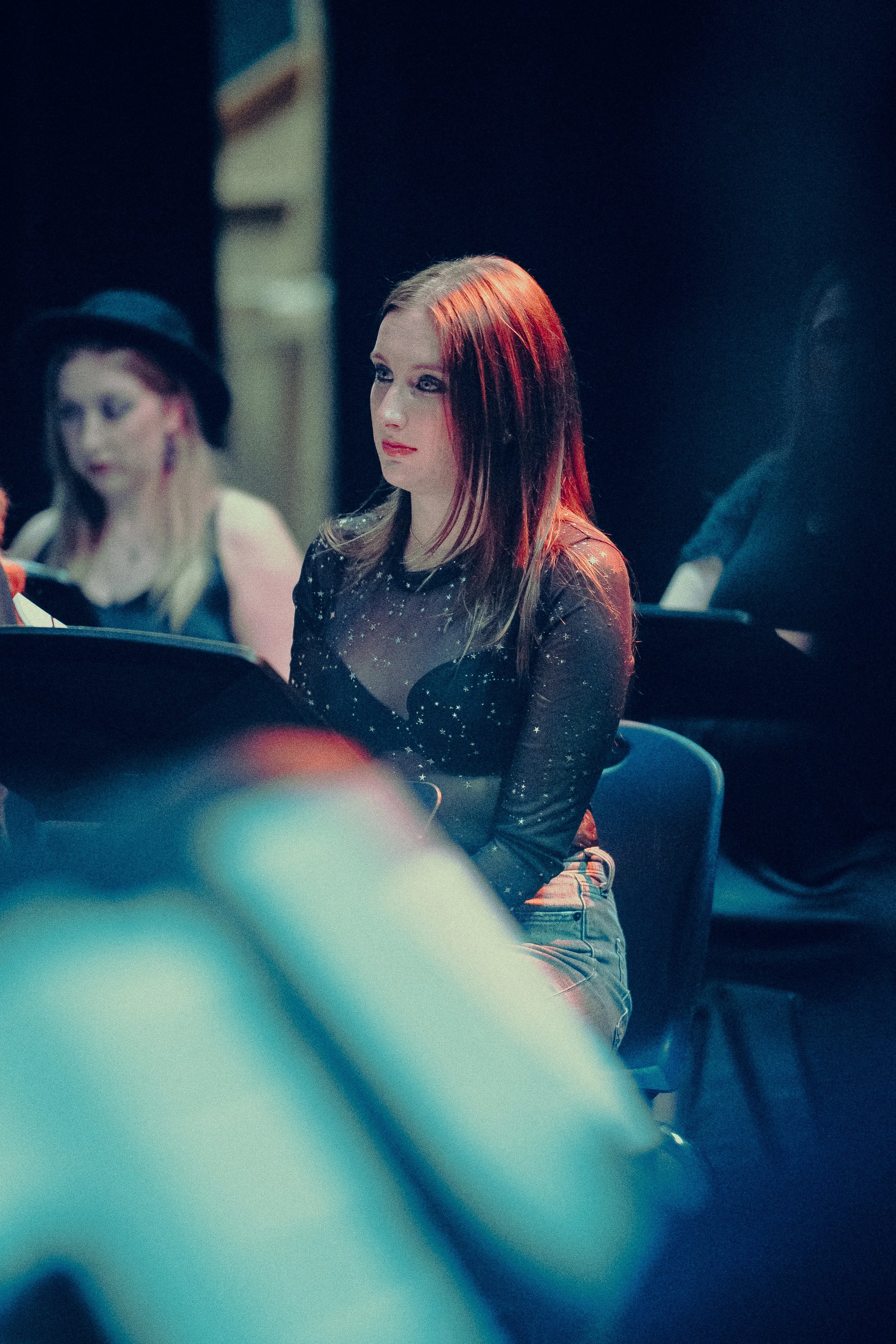 A woman in a sheer black top sits attentively, possibly at an event or performance, with others in the background.
