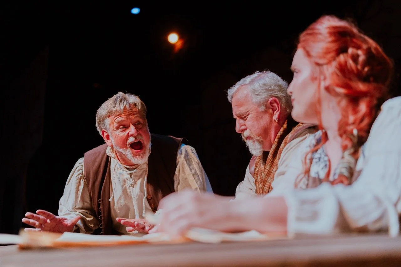 Three people engaged in a theatrical performance, with one passionately gesturing while speaking. The scene is dimly lit, suggesting a dramatic setting.