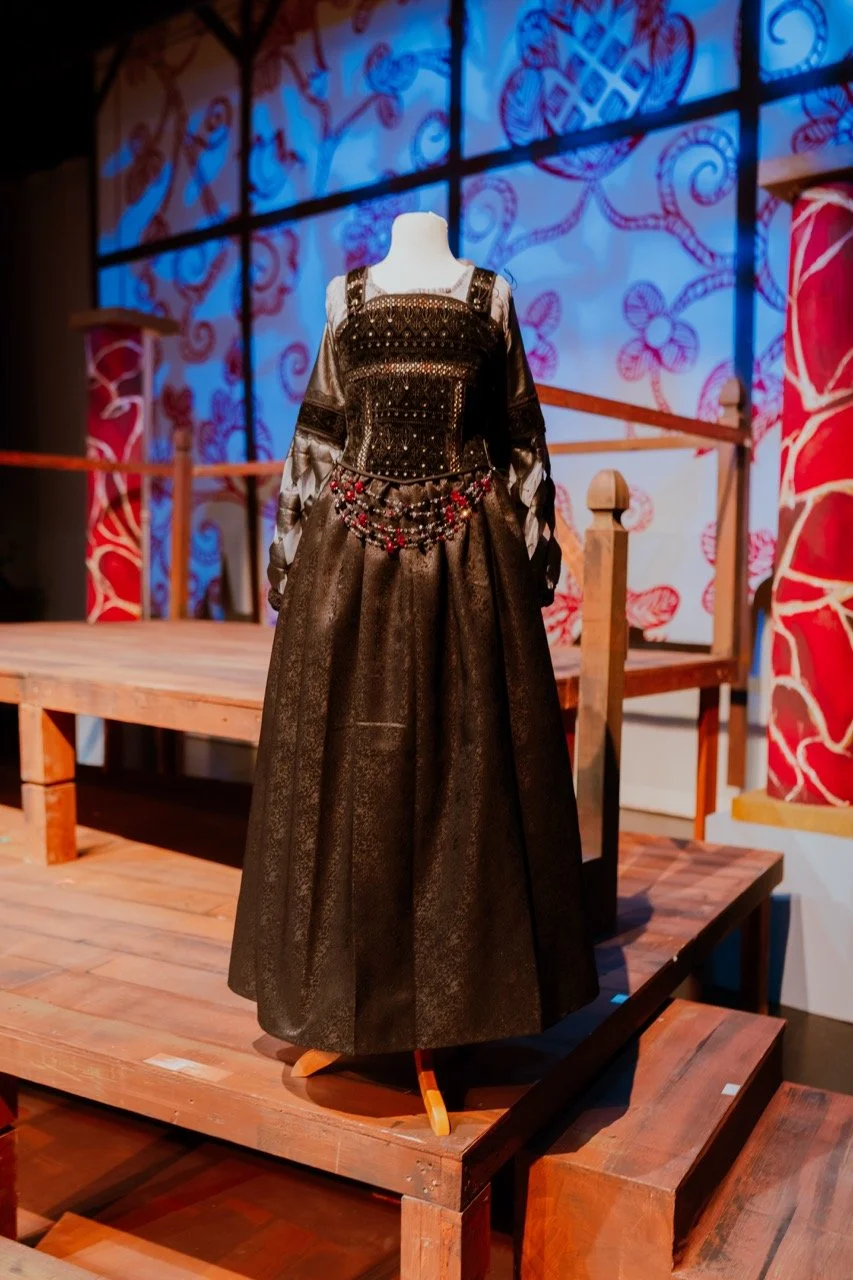 Mannequin displaying an elegant black medieval-style gown with intricate patterns and embellishments on a wooden platform in a decorated room.