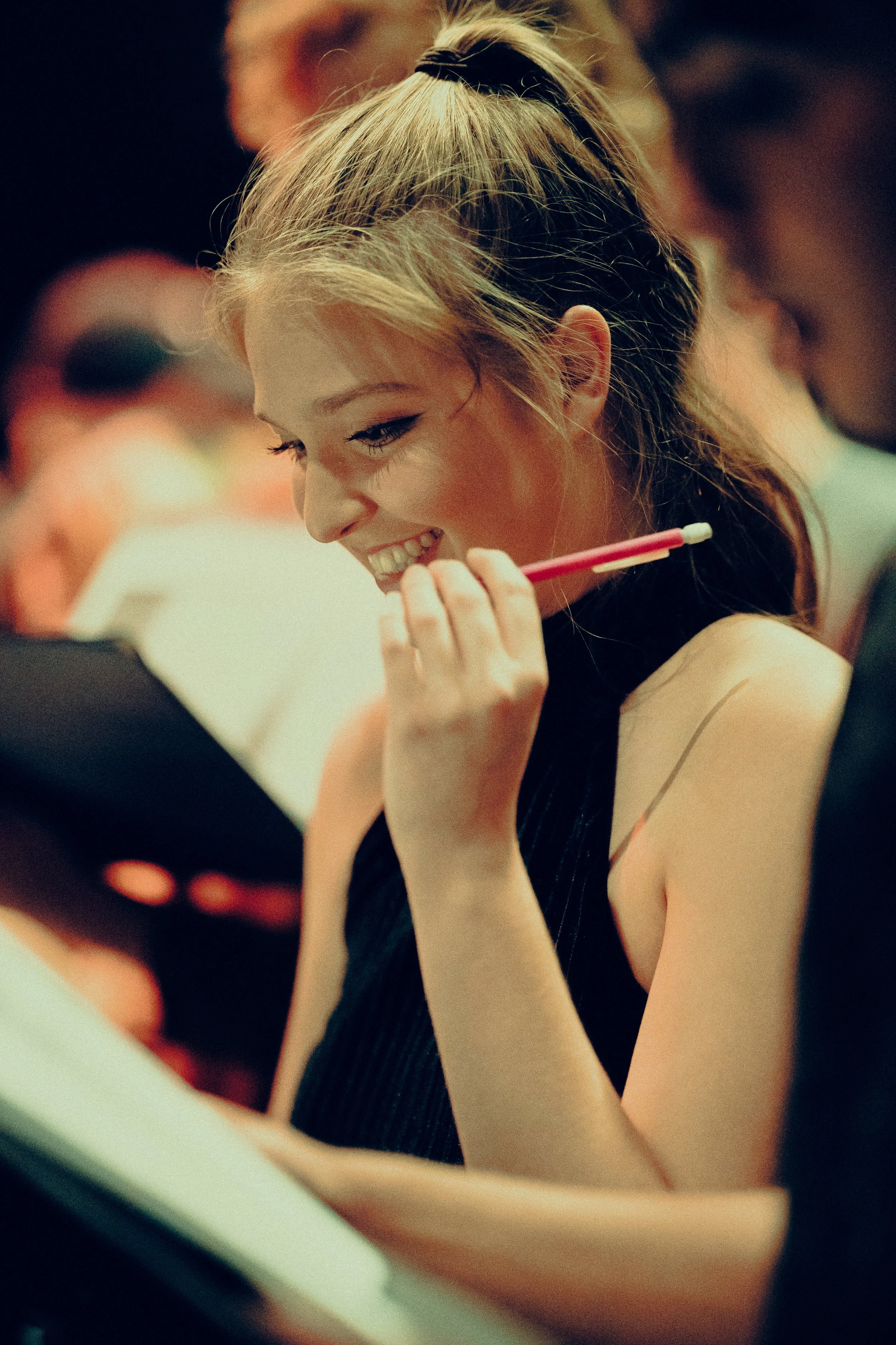 Young woman smiling, holding a pencil, and looking at a paper in a dimly lit setting.