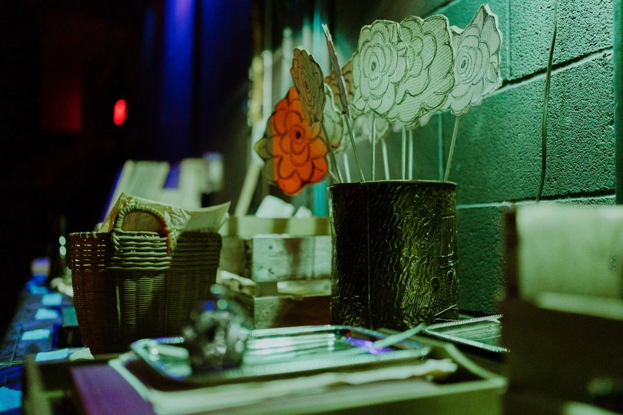Dimly lit scene with hand-drawn flower props in a metal container, wicker basket with envelopes, blurred background.