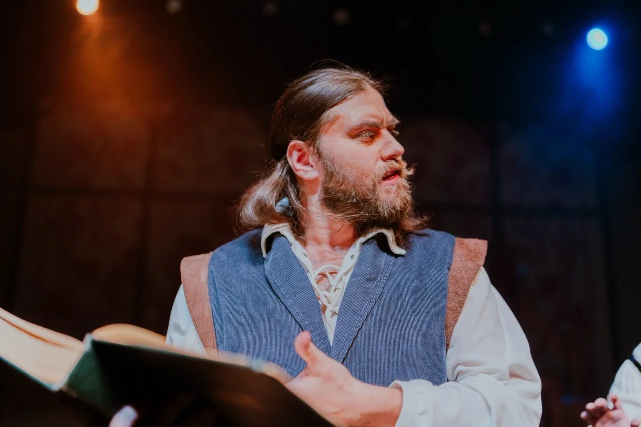 Man in period costume holding a book and looking to the side on stage
