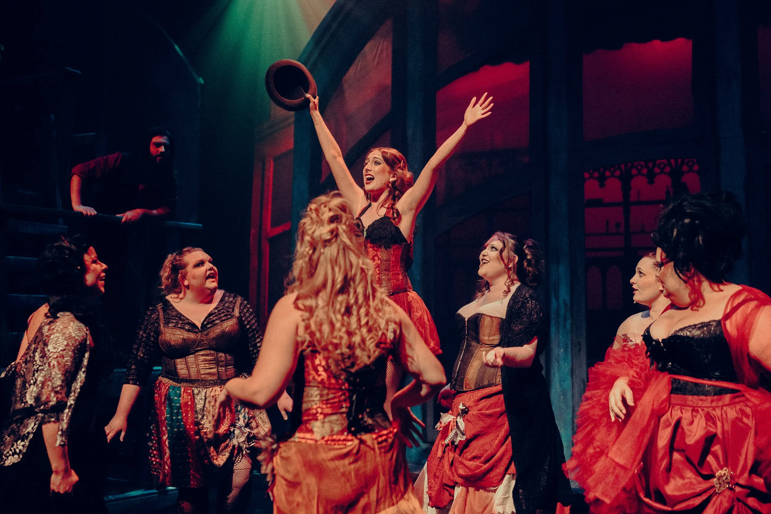 Group of performers in a theatrical scene wearing burlesque-style costumes with black and red attire, one person raising a hat triumphantly under dramatic red and green lighting.