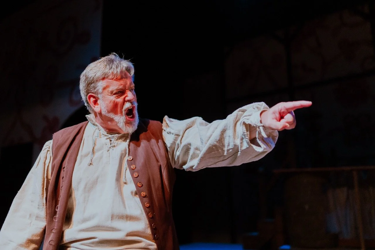 A person in period costume on stage, pointing and appearing to shout during a theatrical performance.