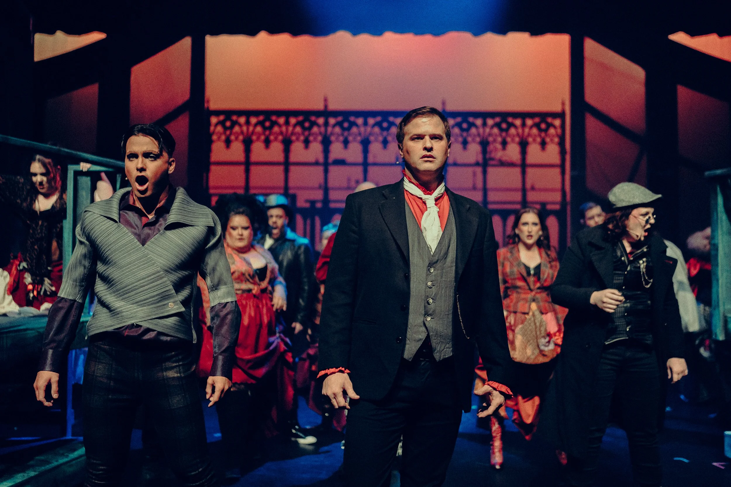 A group of actors on stage performing a dramatic scene in period costumes, featuring two men at the forefront with serious expressions.