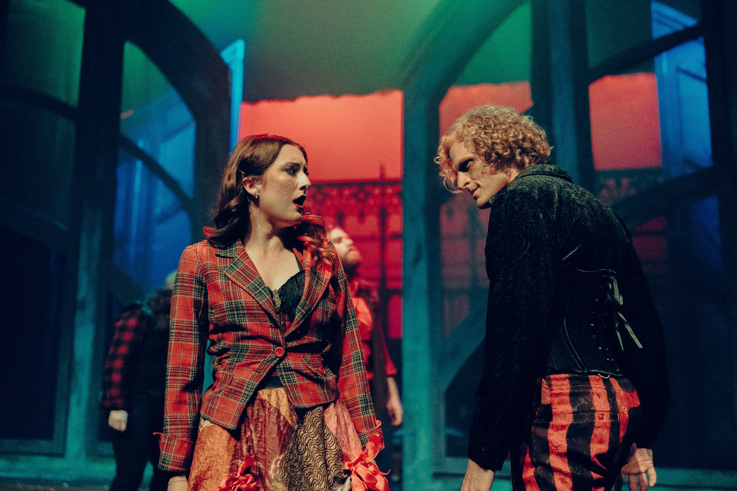 Two actors in colorful, theatrical costumes performing on stage with dramatic lighting and set pieces.