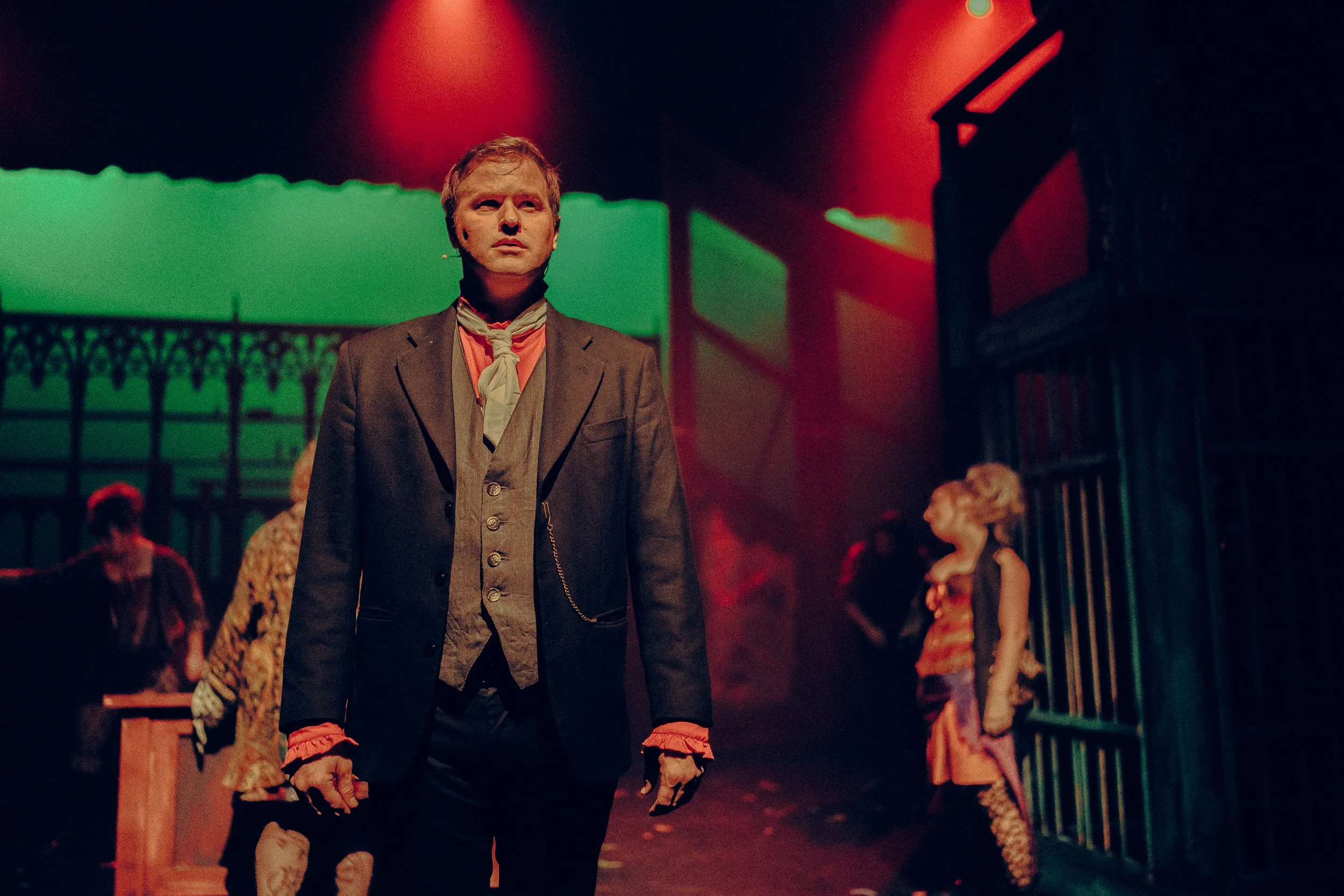 Stage performance featuring man in Victorian-style suit with scar on face under dramatic red and green lighting, background actors and set suggesting period drama.